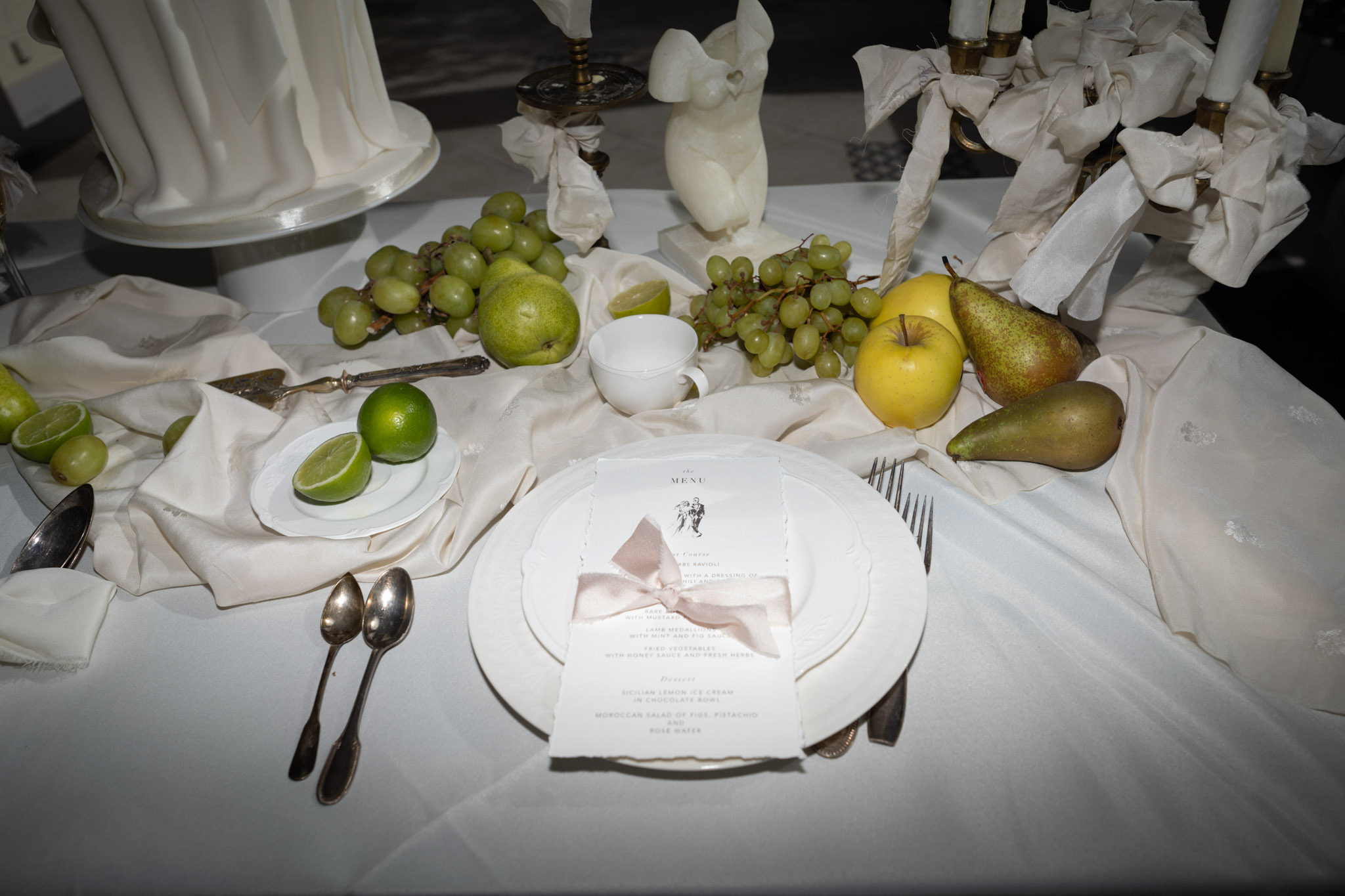 Reception place setting with stacked white plates, menu card tied with pink ribbon, and scattered fruit
