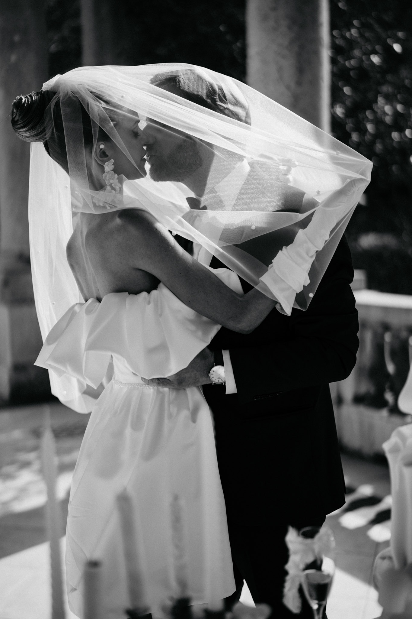 Bride in short white dress and dotted veil kissing groom in dark suit at outdoor ceremony space in B&W