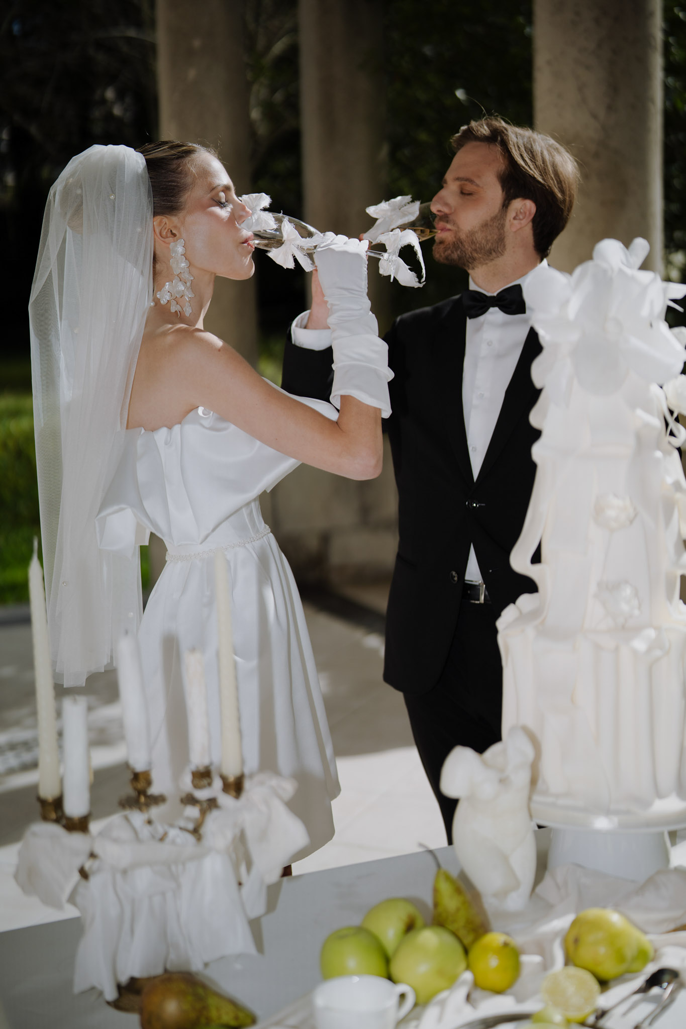 A bride and groom drink champagne together from glasses decorated with white feathers in what appears to be a champagne toast moment, set outdoors near a colonnaded structure. The bride wears a short strapless white satin dress with ruffle tiers and bow details, long white satin gloves, a cathedral-length veil, and oversized white floral drop earrings; the groom wears a black tuxedo with a black bow tie. In the foreground, a tall multi-tiered white wedding cake decorated with large white ruffled sugar flowers is partially visible, alongside a candelabra with white taper candles and a bowl of green pears arranged on a white surface. The overall decor palette is entirely white with a modern yet classic formal styling, and the portrait-style shot frames the couple from roughly mid-torso up.