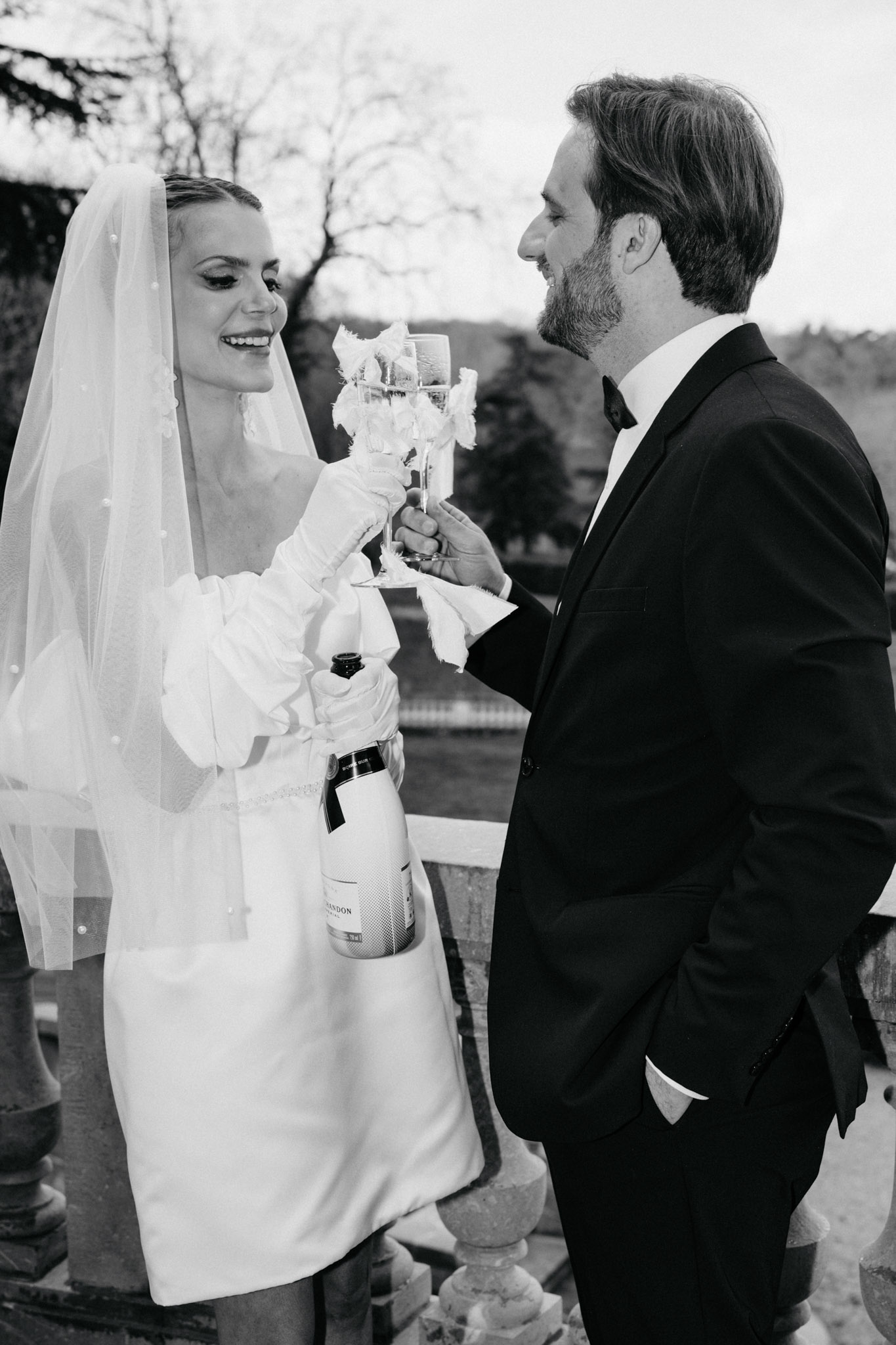 Black-and-white portrait of bride in mini dress with puffed sleeves and groom in tuxedo sharing a champagne toast