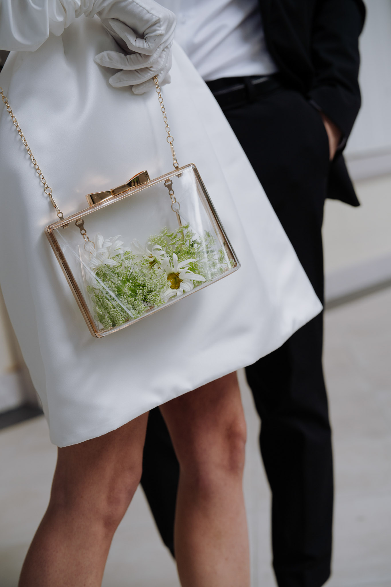 A close-up detail shot focusing on a bride's accessories against her white structured mini dress. She is carrying a clear acrylic box clutch with a gold frame and chain strap, filled with white daisies and green Queen Anne's lace. Her hand is adorned with a white satin or leather glove. Partially visible in the background is a groom in a black suit with black trousers. The overall styling is modern and minimalist, with the floral-filled transparent clutch serving as a standout intentional design detail.