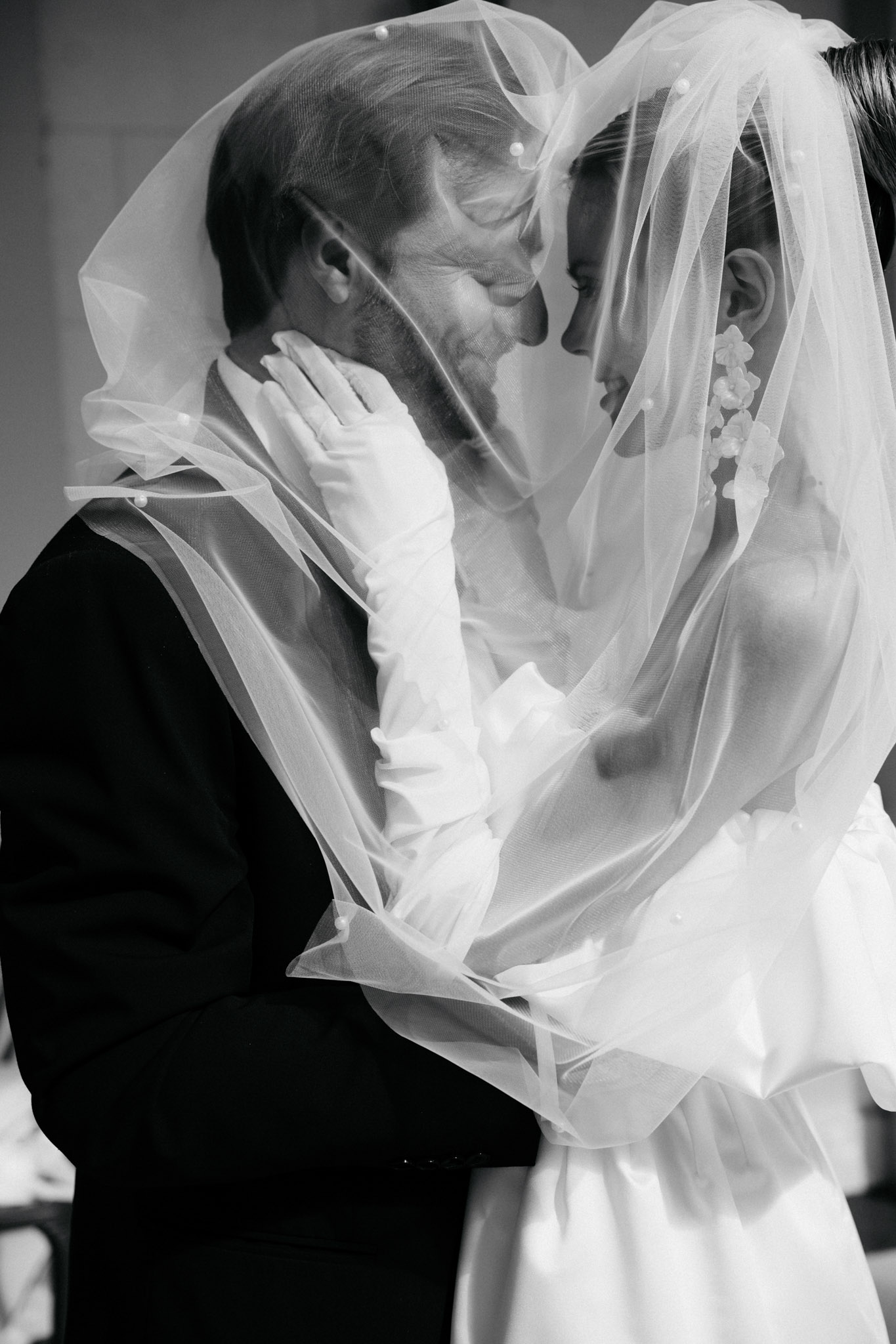 This black-and-white close-up portrait shows a bride and groom sharing an intimate moment with their foreheads touching, both enveloped under the bride's veil. The bride wears elbow-length white satin gloves and has small floral hair accessories visible near her ear; her veil is dotted with pearl-like embellishments. The groom wears a dark suit jacket. The veil drapes over both of them, creating a soft, layered composition with bright whites against deep shadow tones. The image is a tight mid-shot portrait with strong contrast between the sheer fabric and the dark background.