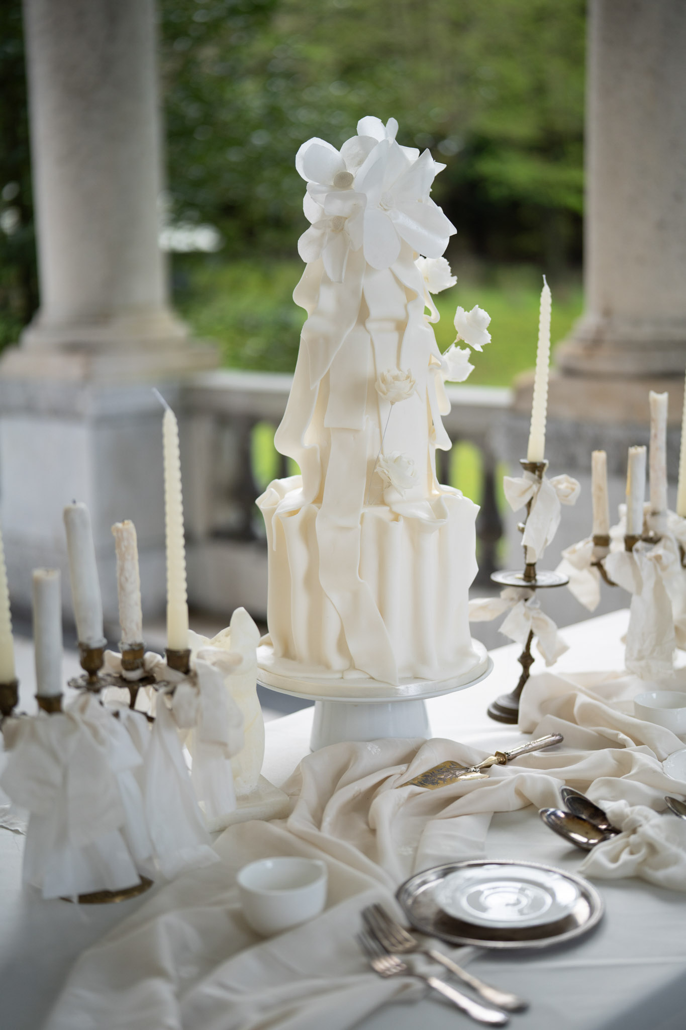 Two-tier ivory wedding cake with draped fondant and sugar flowers on pedestal, brass candelabras alongside