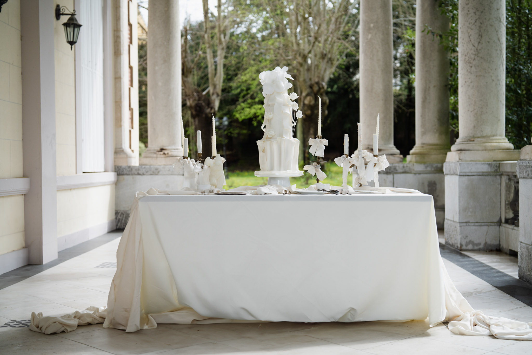 A styled wedding tablescape displayed on a covered outdoor colonnade or portico, framed by tall white stone columns. The rectangular table is draped in an ivory linen cloth that pools deliberately on the tiled floor at both corners. Centered on the table is a tall all-white multi-tiered wedding cake with vertical ribbed detailing and oversized white sugar floral decorations cascading from the top. The table is flanked symmetrically by brass candelabras holding white taper candles, each decorated with white ribbon bows. Place settings with grey charger plates, silverware, and white folded napkins are arranged on either side of the cake. The overall decor palette is a strict all-white and ivory monochrome scheme with a classic, formal styling approach. Wide shot, slightly low angle.