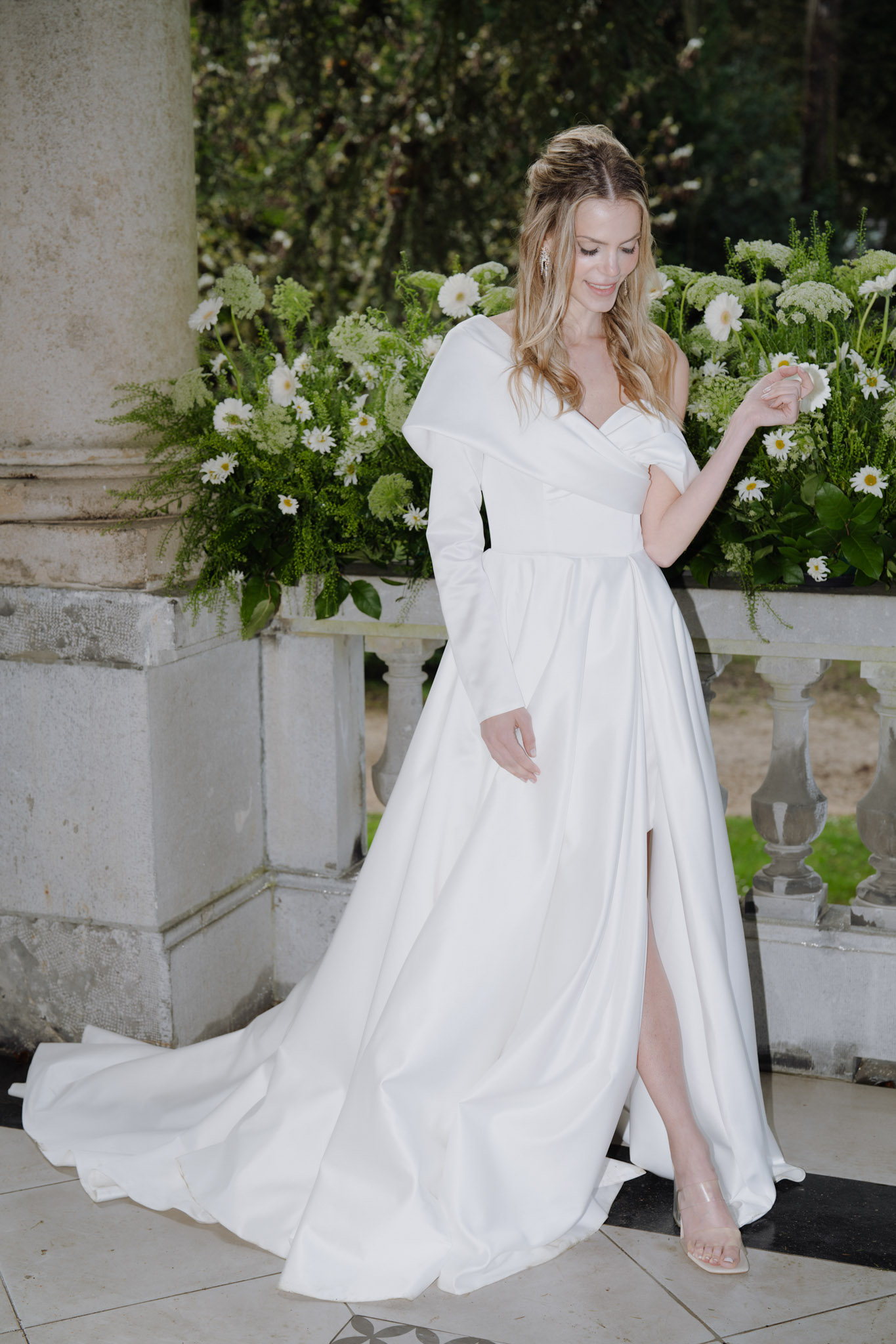 Bride in ivory asymmetric one-sleeve gown posing on stone terrace with white daisy and green floral arrangements behind