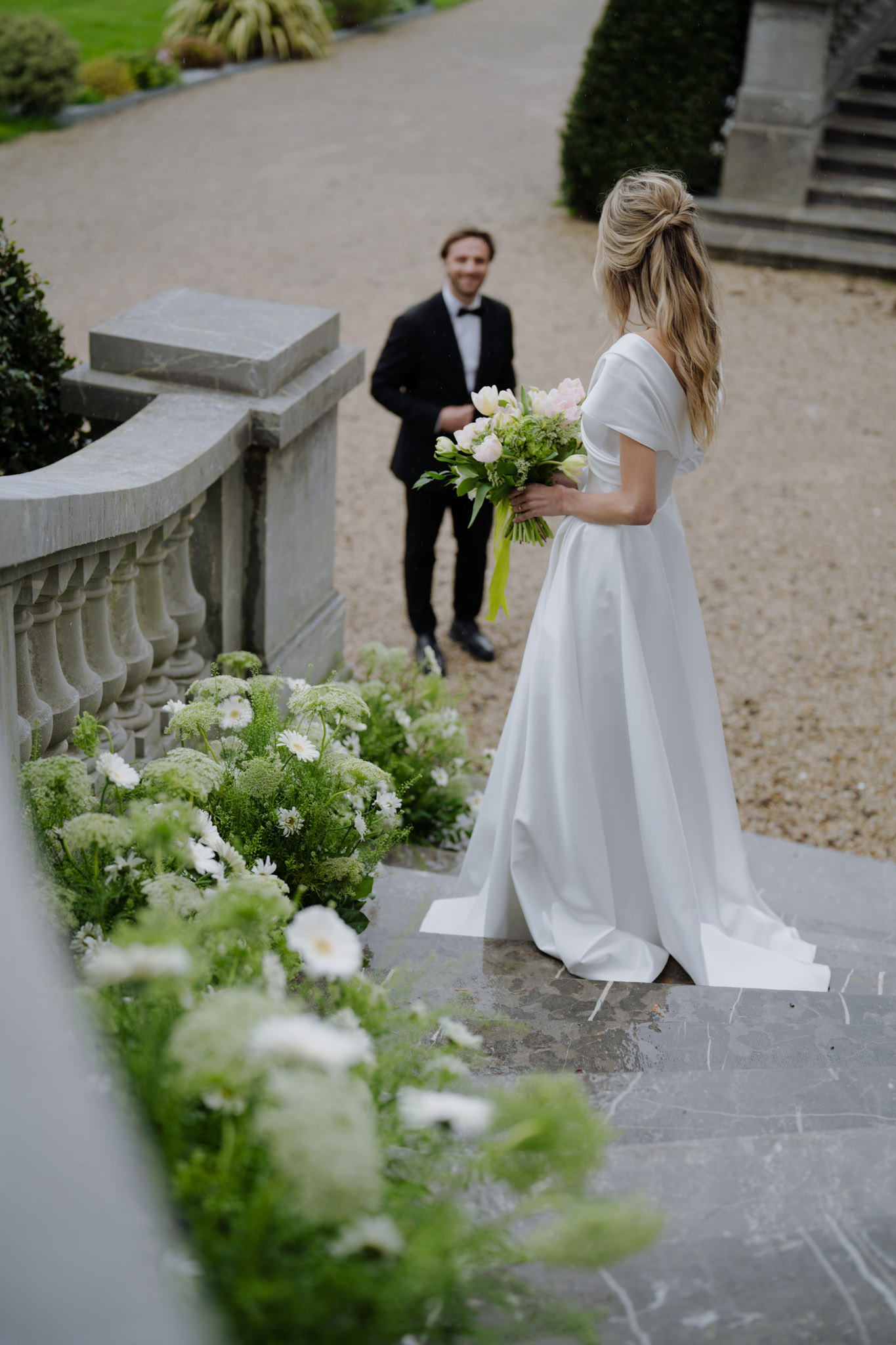 Bride with blush peony bouquet on stone terrace groom on gravel path below with daisy-lined staircase at chateau