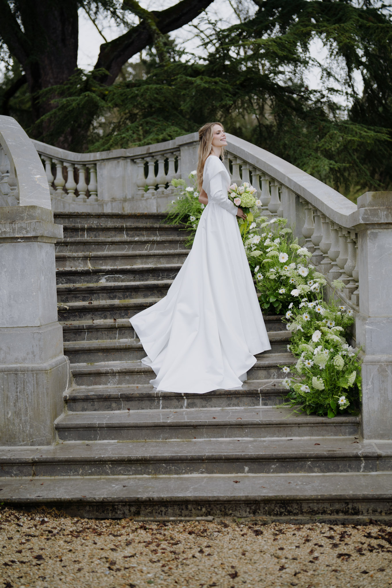 Bride in long-sleeve white gown with blush bouquet on stone staircase with cascading white daisy installation