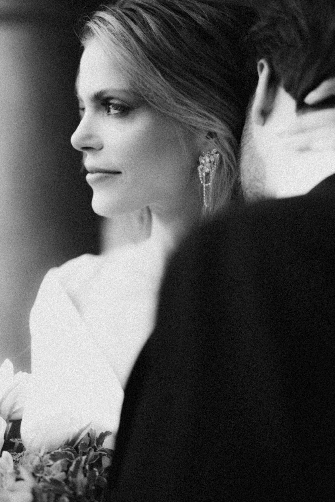Black and white close-up of bride with crystal earrings and bouquet framed by groom's shoulder