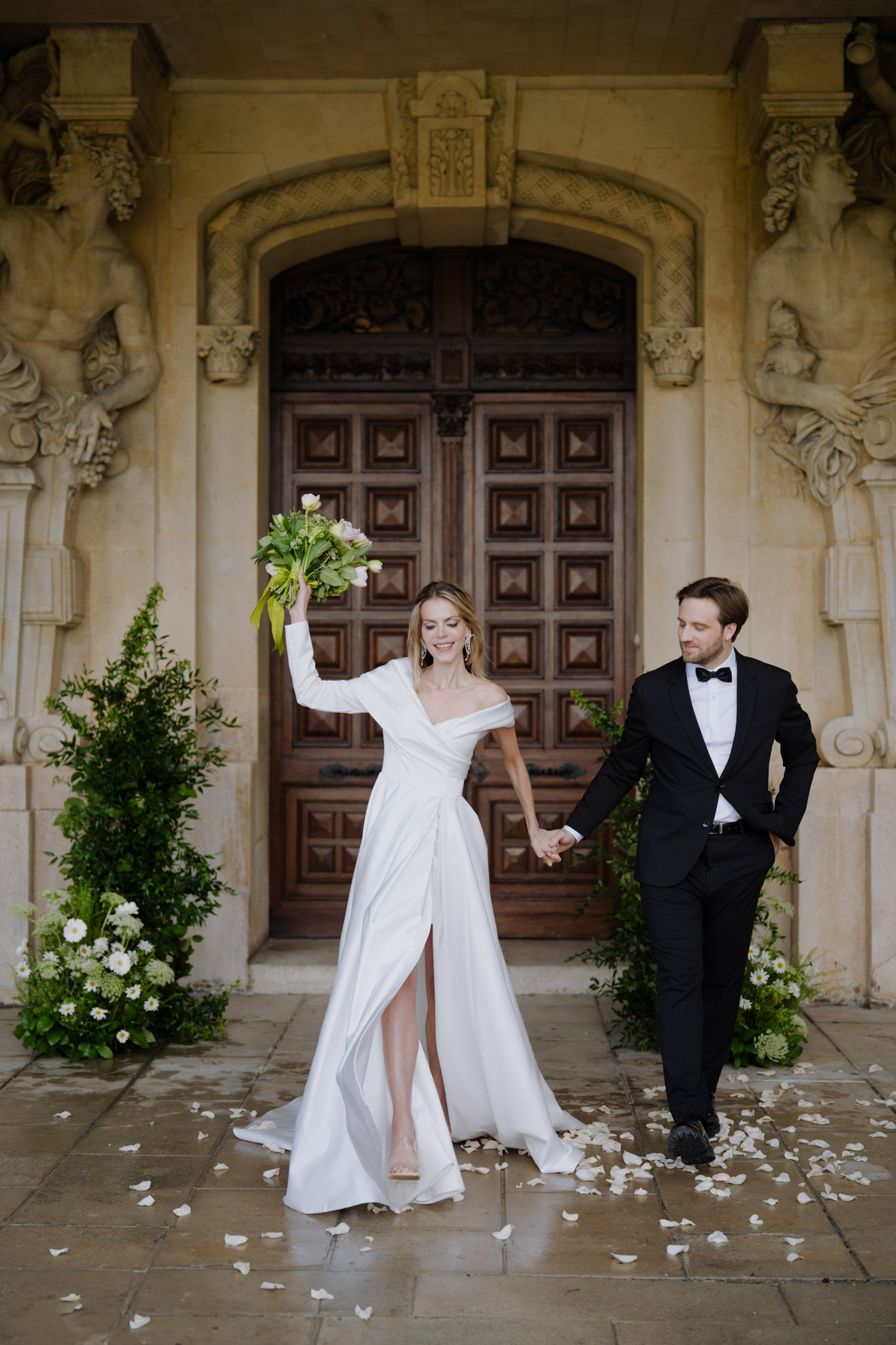 Bride in one-shoulder gown and groom in tuxedo walking past ornate stone doorway with scattered petals