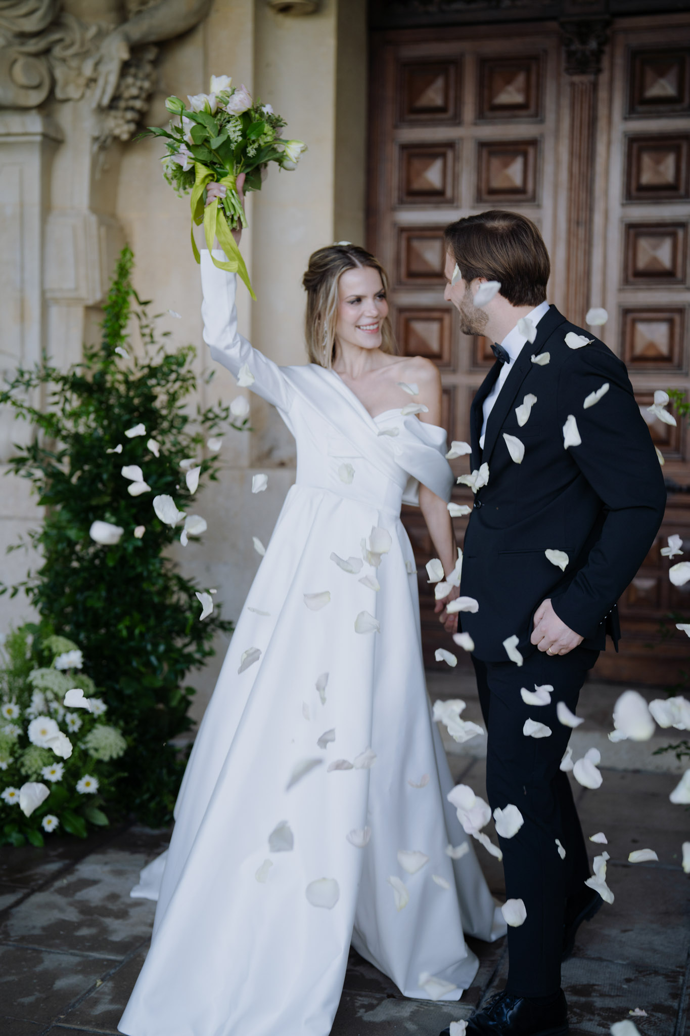 Couple holds hands in rose petal exit at carved stone chateau doorway with bride's bouquet raised high