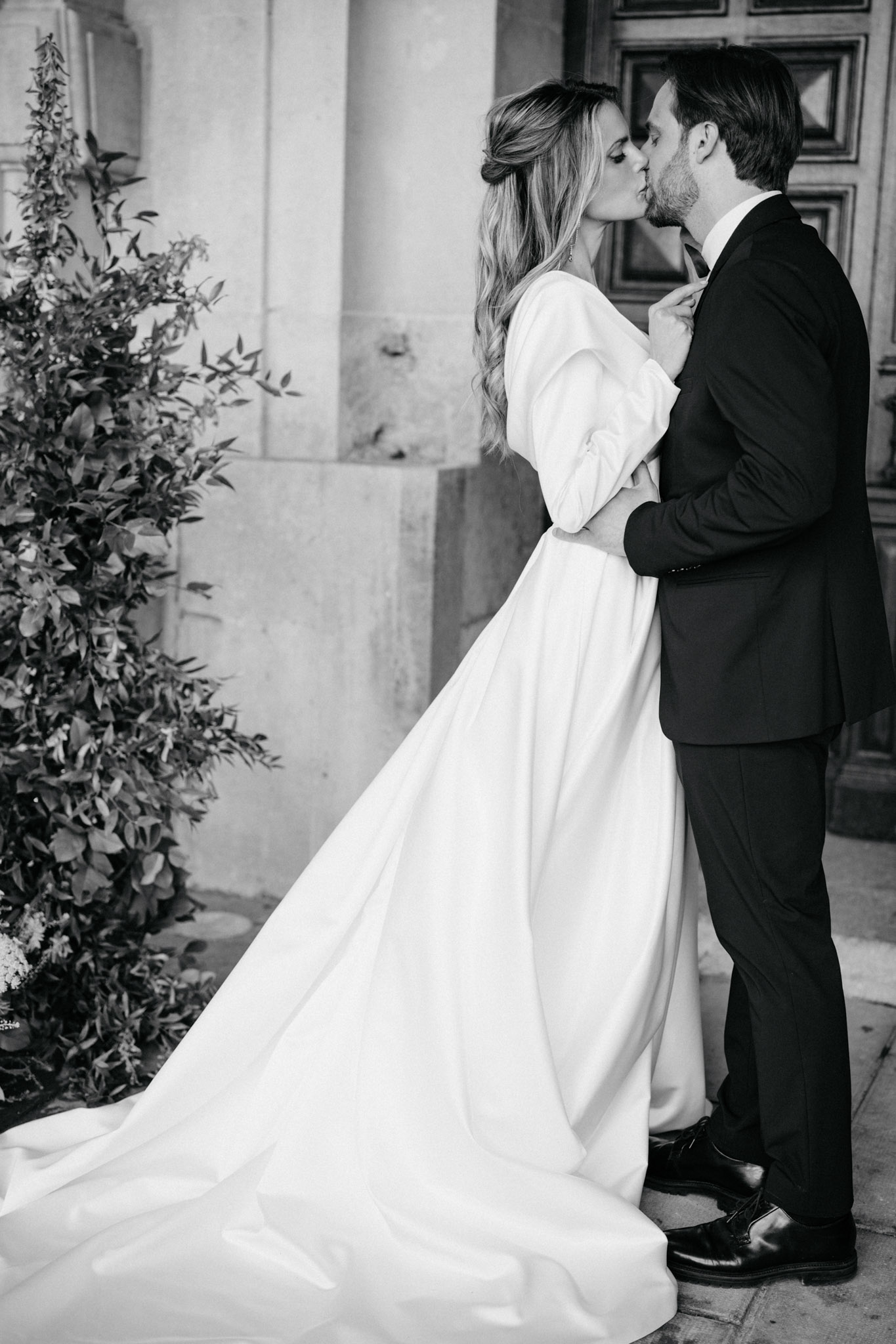 This is a black-and-white couple portrait taken outdoors against the stone facade of a formal building, with a large ornate wooden door visible in the background. The bride and groom are kissing, with the groom's arms wrapped around the bride's waist and her hand resting on his chest. The bride wears a full-skirted gown with structured puff sleeves and a long flowing train, styled in a clean, modern silhouette; her hair is worn down in loose waves with a partial updo. The groom wears a dark suit with a bow tie. A tall foliage arrangement featuring leafy branches is positioned to the left of the couple. The image is a full-length portrait shot with high contrast and rich dark tones, emphasizing the clean lines of the dress against the architectural backdrop.