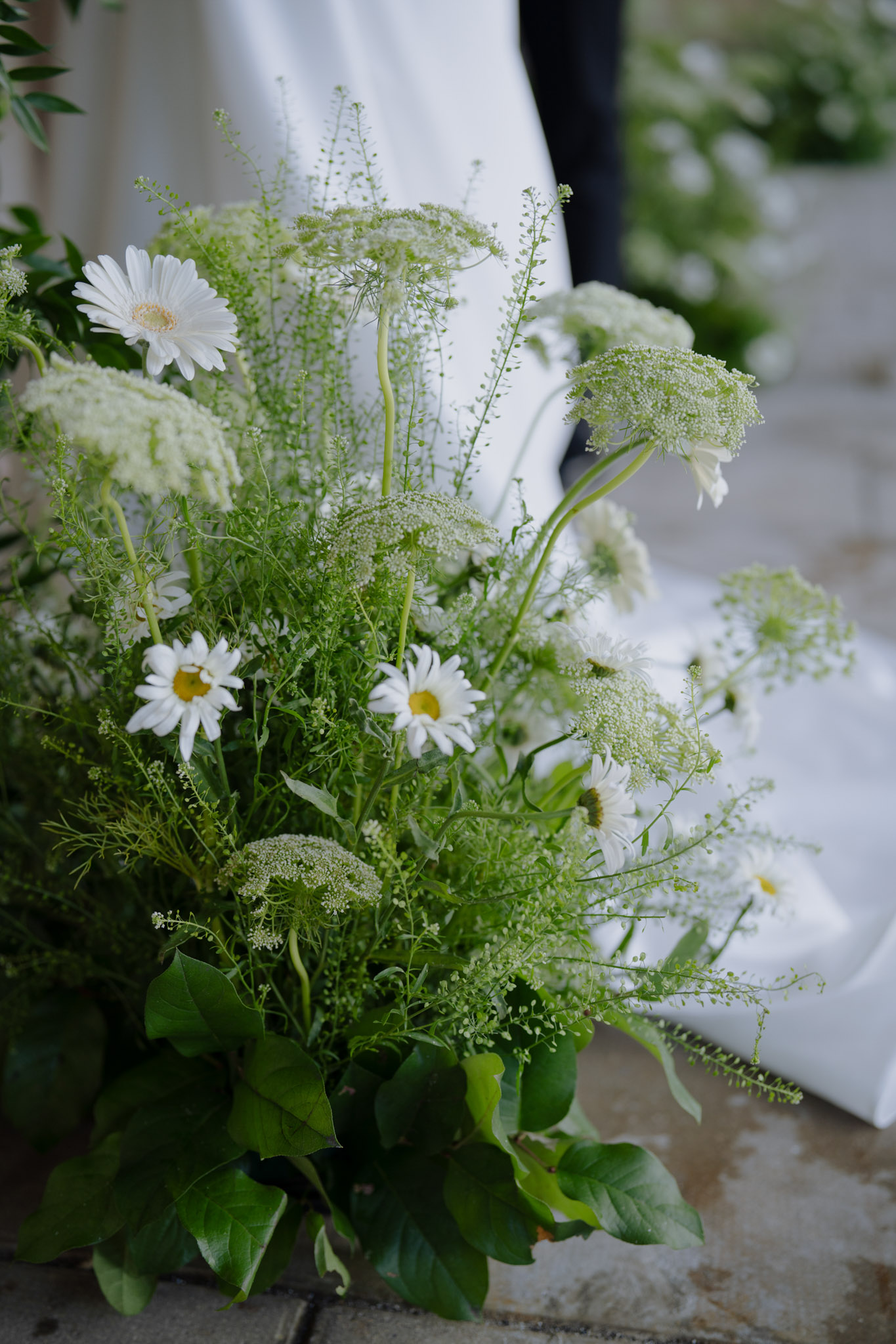 Wild garden-style ceremony floral arrangement of white daisies, Queen Anne's lace, and green foliage on stone floor