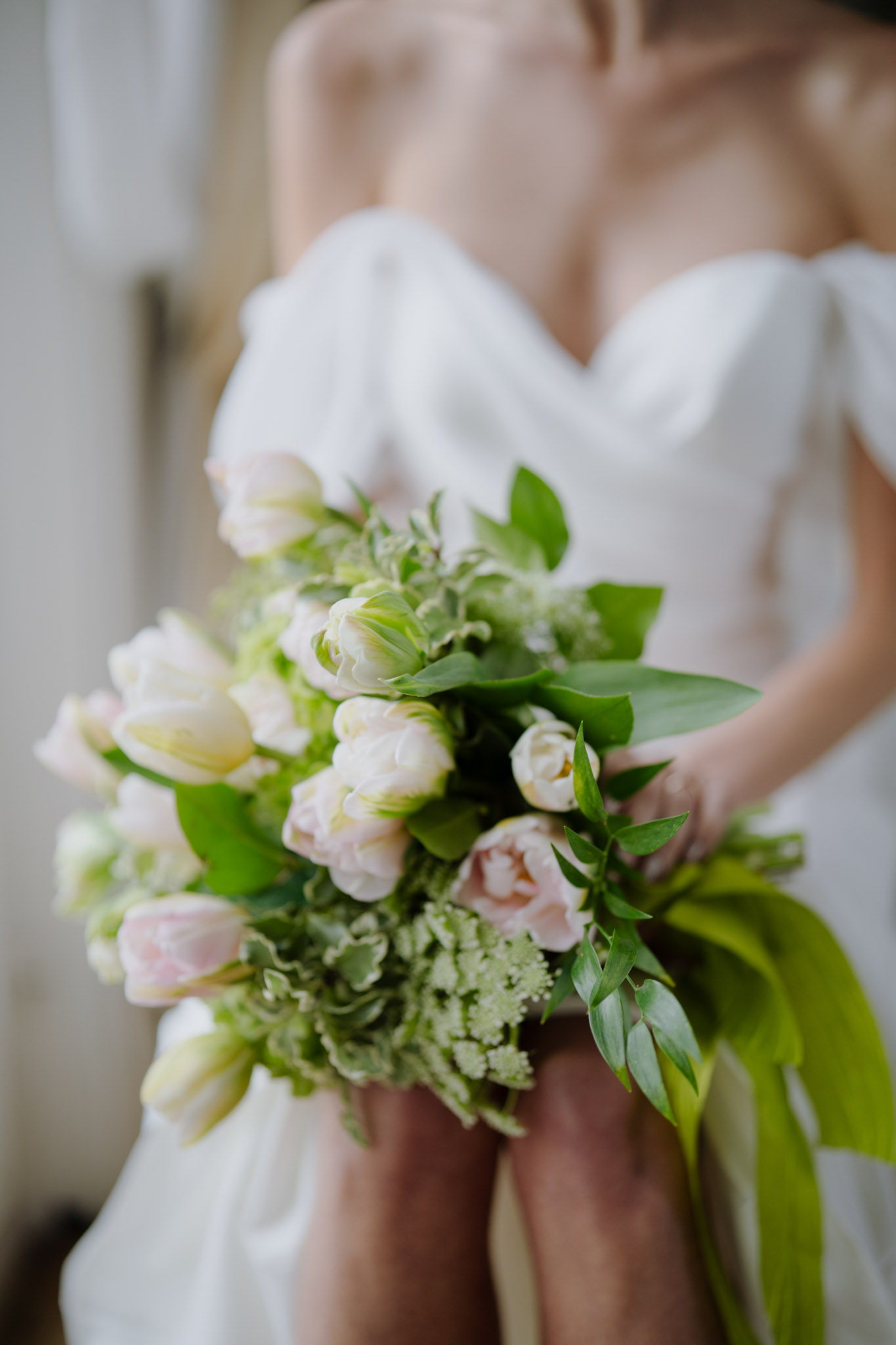 Bride holding bouquet of blush tulips, cream garden roses, white filler flowers, and trailing green foliage with green ribbon