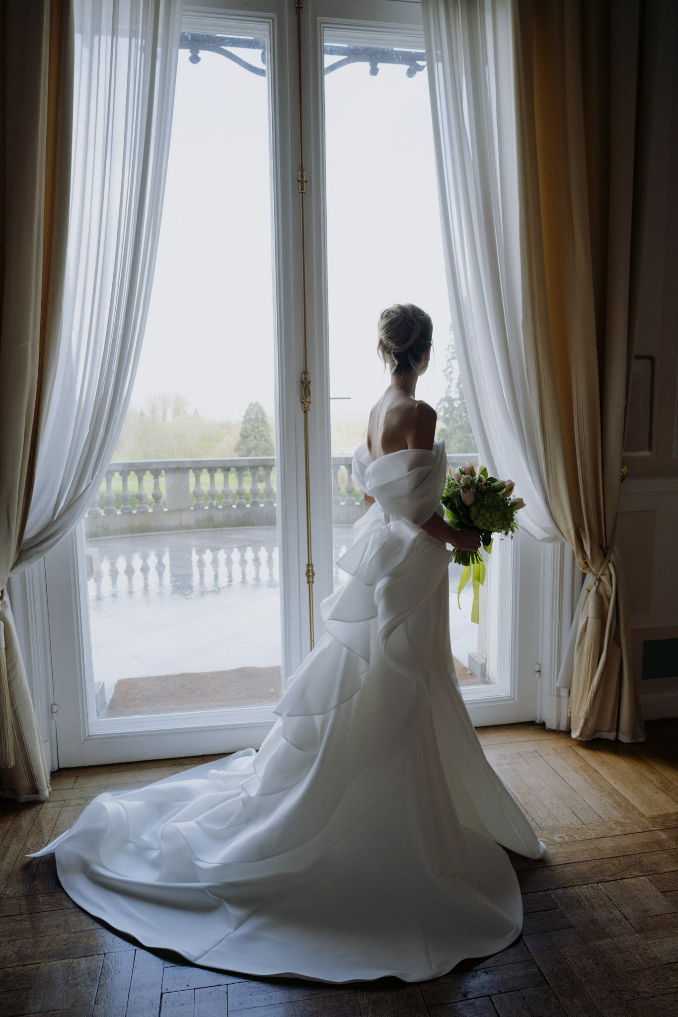 A bridal portrait taken indoors in what appears to be a château or manor house, with the bride standing with her back to the camera at a set of tall French doors. She wears a white off-the-shoulder gown with a structured oversized bow at the back and a long cathedral train that spreads across the herringbone parquet floor. Her hair is styled in an updo. She holds a compact bouquet of soft pink tulips and green hydrangea with a bright green ribbon wrap. Floor-length cream and white curtains frame the doors, and through the glass a stone balustrade terrace is visible. The composition is a full-length portrait shot from behind, using natural backlight from the doors to silhouette the dress and train.