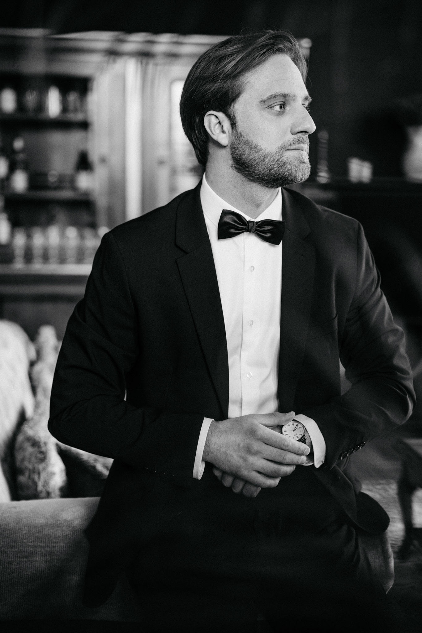 Black and white portrait of groom seated indoors holding pocket watch wearing tuxedo and bow tie