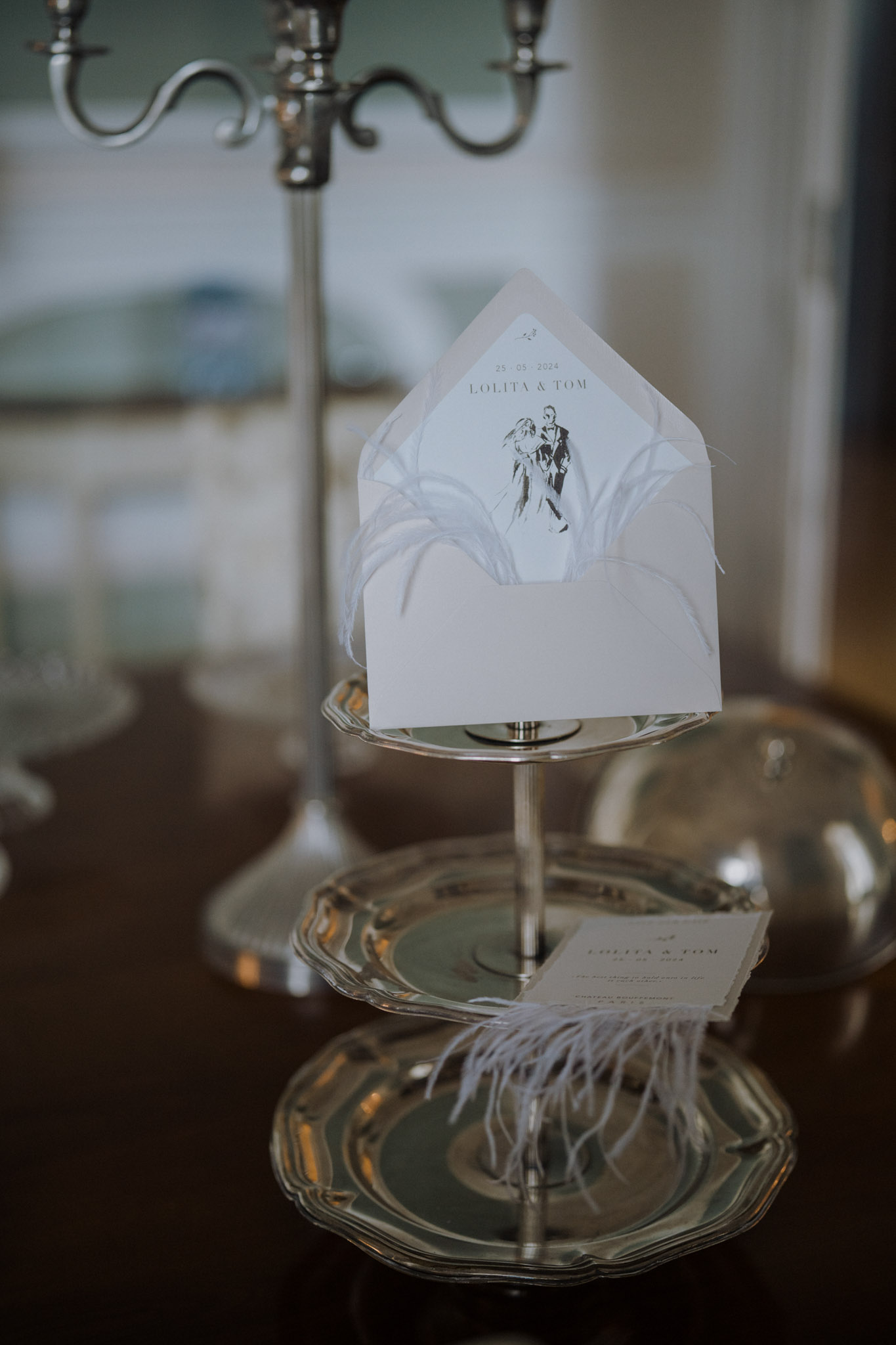 A close-up detail shot of wedding stationery displayed on a two-tiered silver tray stand placed on a dark wooden surface. The upper tier holds an open white envelope featuring a custom illustrated liner with a hand-drawn sketch of a dancing couple and the text '25 · 05 · 2024 / LOLITA & TOM'; white feather trim is tucked into the envelope for decoration. The lower tier holds a matching flat invitation card, also reading 'Lolita & Tom', tied with white feather embellishments. A silver candelabra is partially visible and softly out of focus in the background. The stationery suite follows a classic, monochrome design palette with illustrative custom artwork and feather styling details. The overall composition is an indoor styled detail shot with shallow depth of field.
