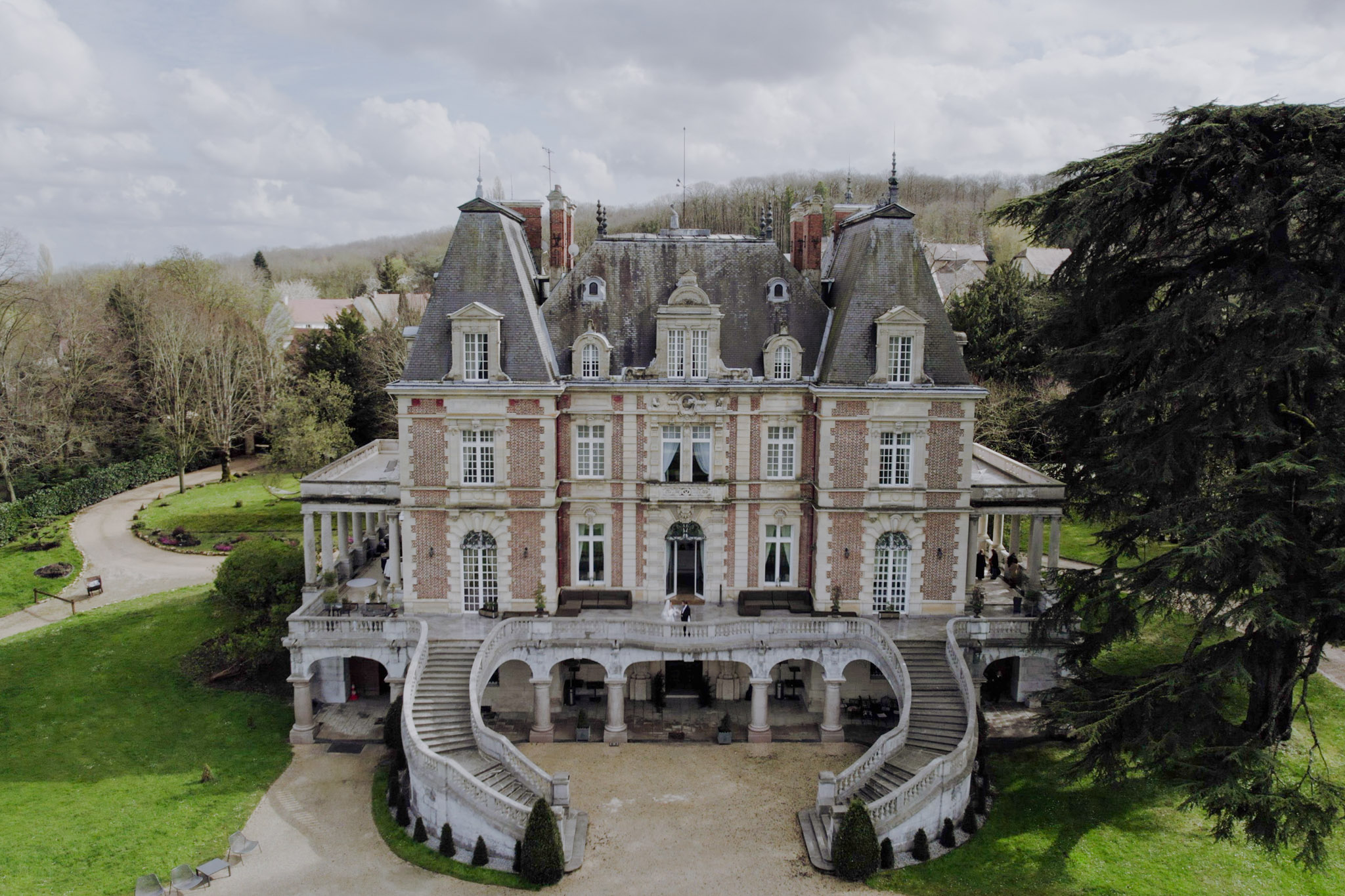 An aerial drone shot captures the full façade of a French château wedding venue, featuring red brick and cream stone architecture with a slate mansard roof, dormer windows, and ornate corner towers. The château's most distinctive feature is its grand double horseshoe staircase in white stone, curving symmetrically down from a wide terrace supported by arched colonnades. Two figures who appear to be a couple in wedding attire — one in white — are visible standing at the top center of the staircase on the terrace, and a small group of guests can be seen on the right-hand terrace colonnade. The surrounding grounds include a gravel driveway, trimmed box hedges, and mature trees including a large cedar to the right. Potential venue feature image.