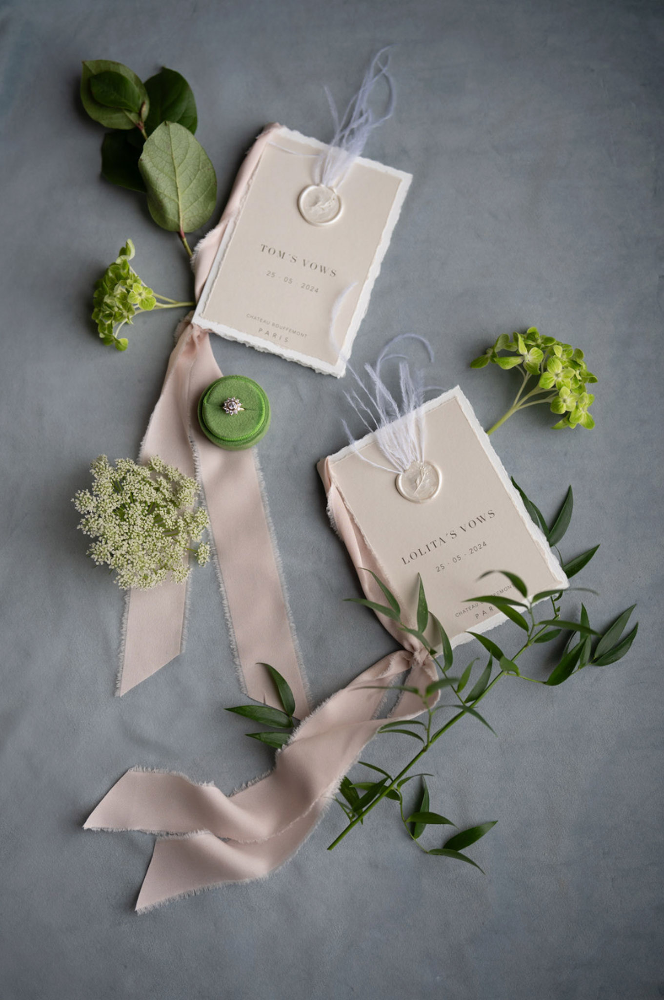 A flat lay detail shot of two wedding vow booklets on a muted blue-grey surface. The booklets are printed on cream deckle-edged paper with minimal serif typography, labeled 'Tom's Vows' and 'Lolita's Vows' dated 25.05.2024 at Château Bouffémont, Paris, each sealed with a cream wax stamp and tied with fine white ribbon or silk thread. A length of frayed-edge blush silk ribbon weaves between the two booklets, and a small green velvet ring box containing a diamond ring rests on top. Greenery sprigs including eucalyptus, viburnum, and what appears to be Queen Anne's lace are scattered around the composition for styling. The overall palette is cream, blush, and green, with a clean modern-classic stationery aesthetic.
