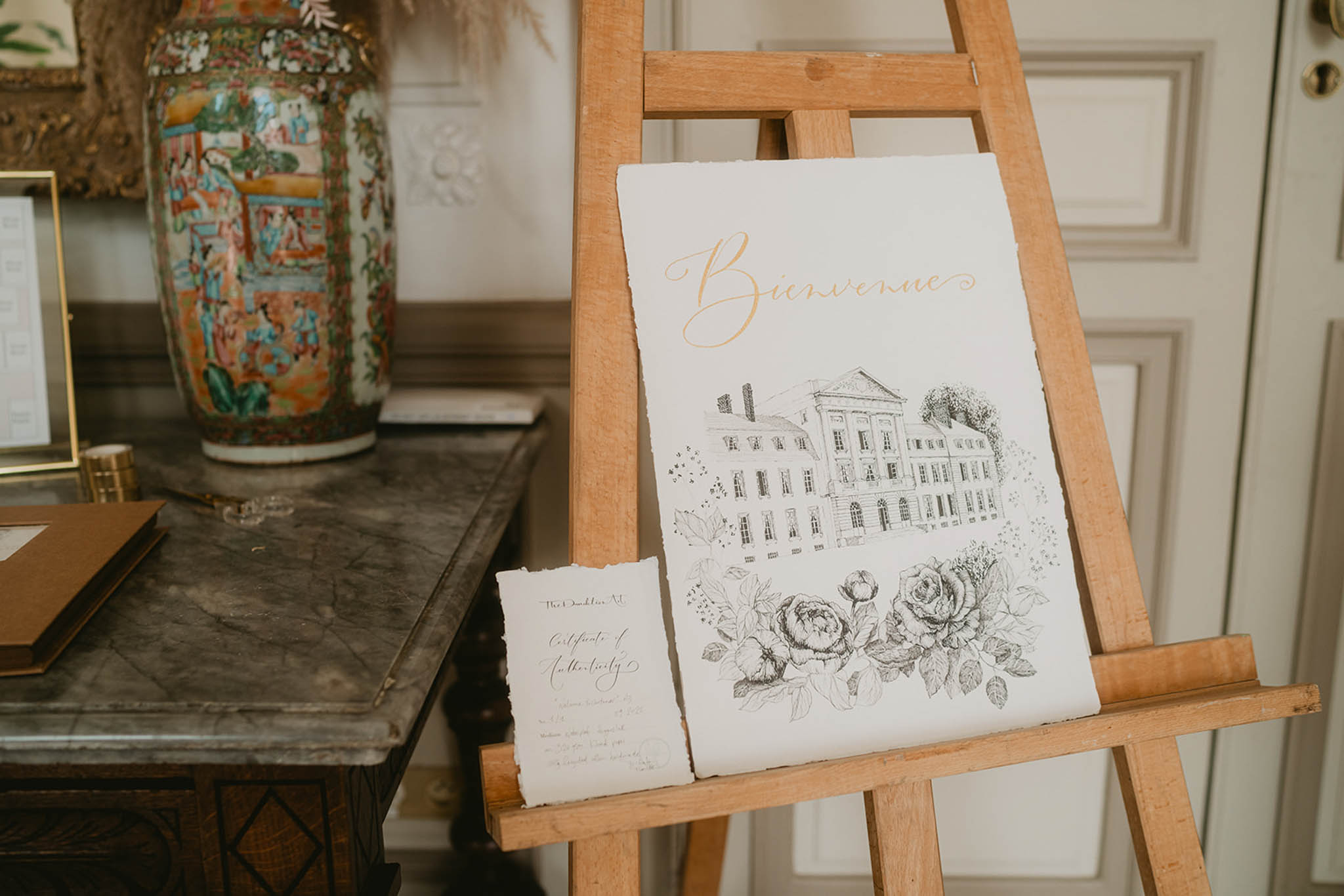 A close-up detail shot of a wedding welcome sign displayed on a natural wood easel, positioned indoors near a panelled door. The sign is printed on deckle-edged white paper and features the word 'Bienvenue' in gold script calligraphy at the top, below which is a hand-drawn black ink illustration of a large classical French château facade surrounded by detailed botanical drawings of roses and peonies. A small certificate of authenticity card leans against the base of the sign on the easel. In the background, a dark antique wooden console table holds a large decorative Chinese famille rose porcelain vase, a gold-framed item, a tan leather guest book, and a small gold object. The overall stationery style is classic and illustrative with a black ink and gold colour palette.