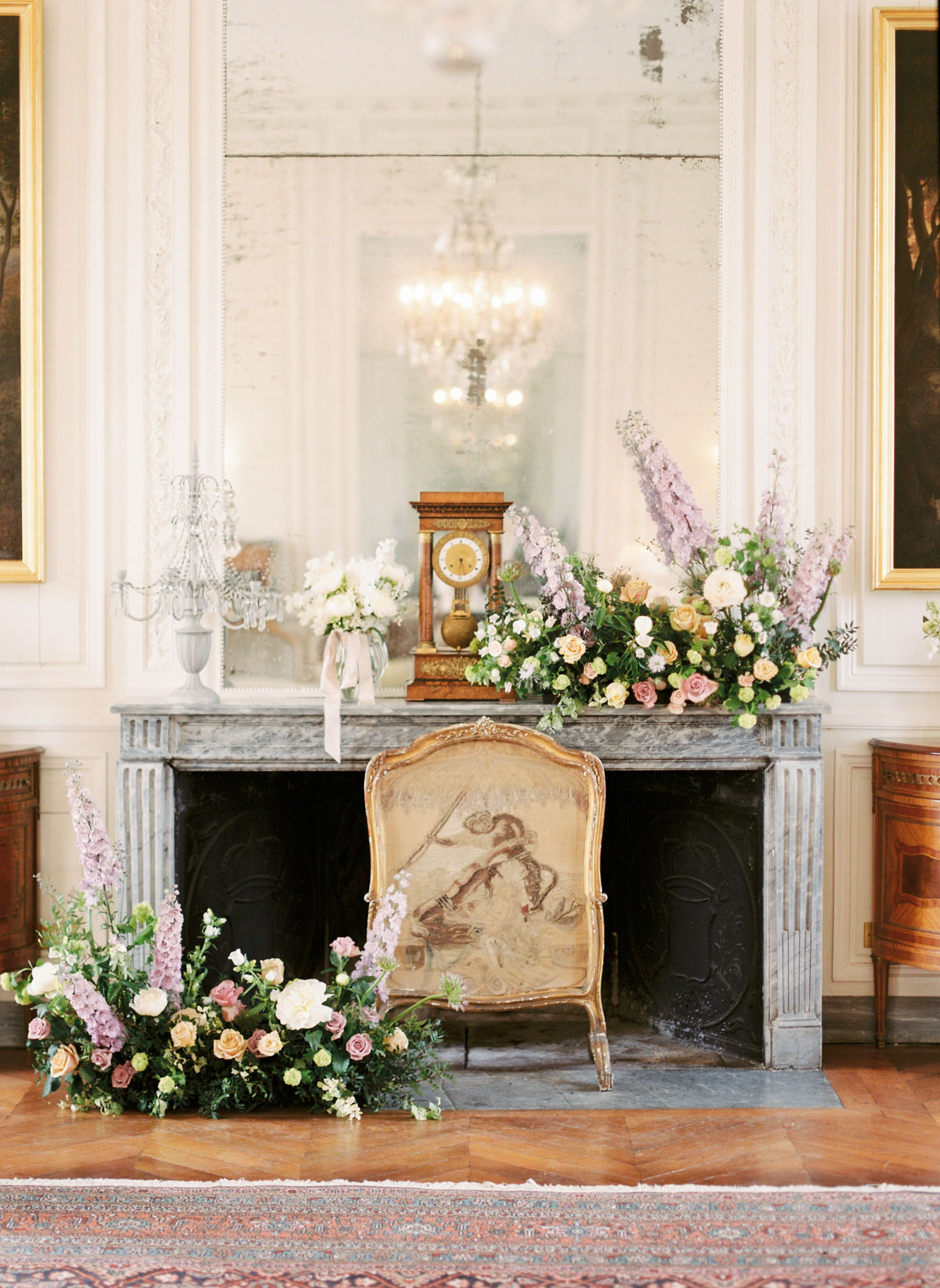 French chateau salon with marble fireplace styled with blush, peach, and lavender floral arrangements on herringbone parquet