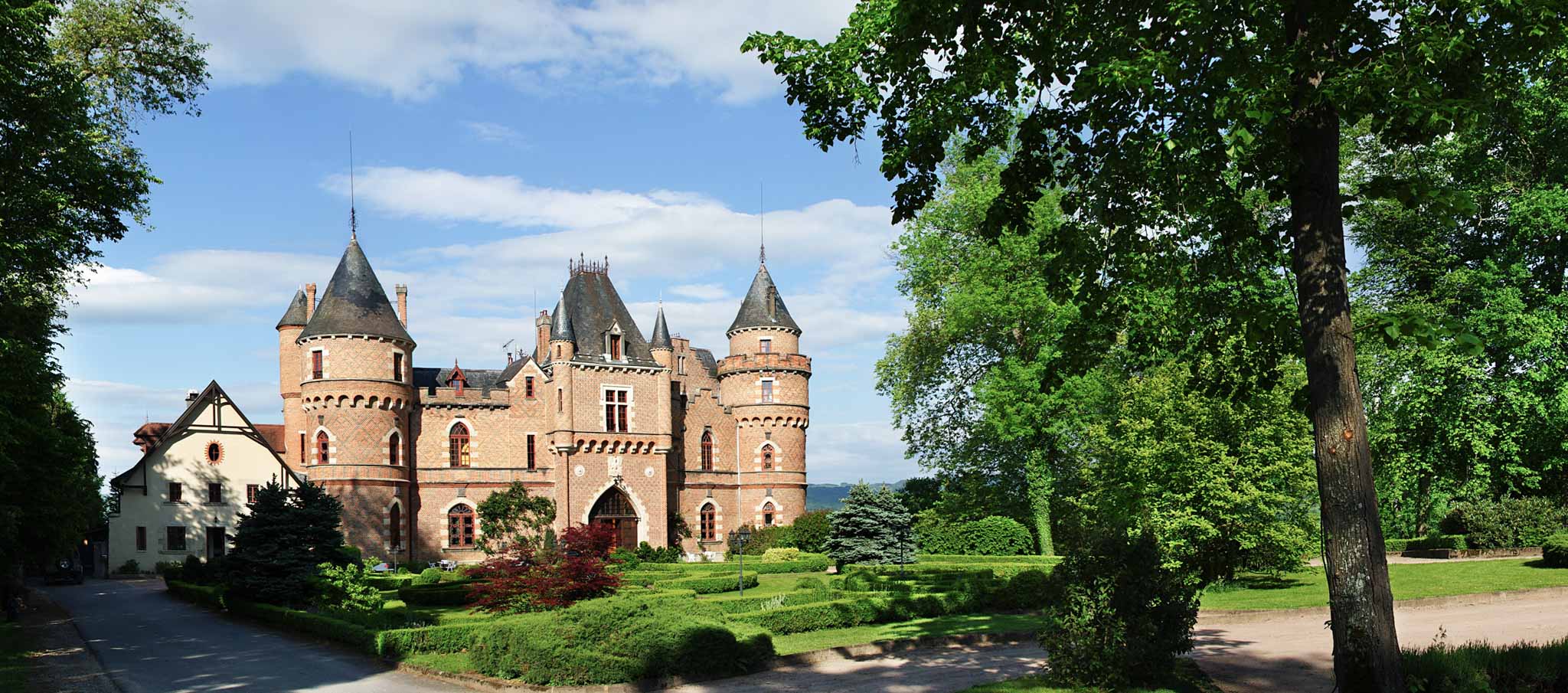 Wide exterior of pink-red brick French chateau with conical turret towers and formal hedgerow gardens