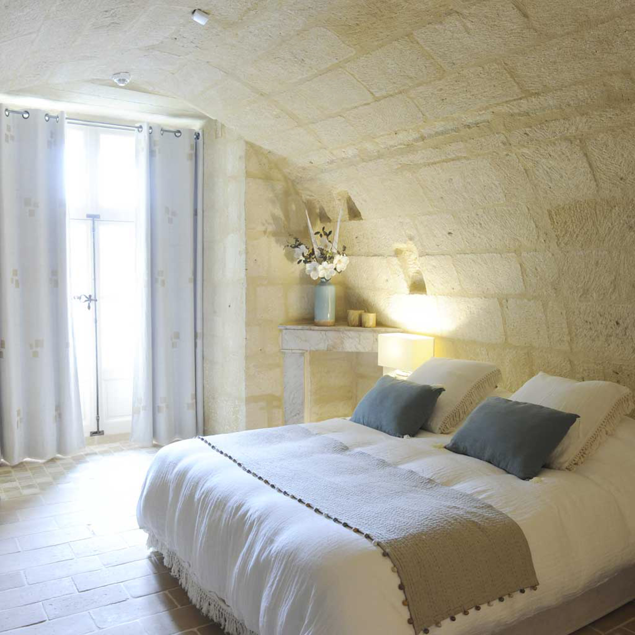 Guest bedroom in French chateau with vaulted stone walls, white linen bed, blue velvet cushions, and stone fireplace