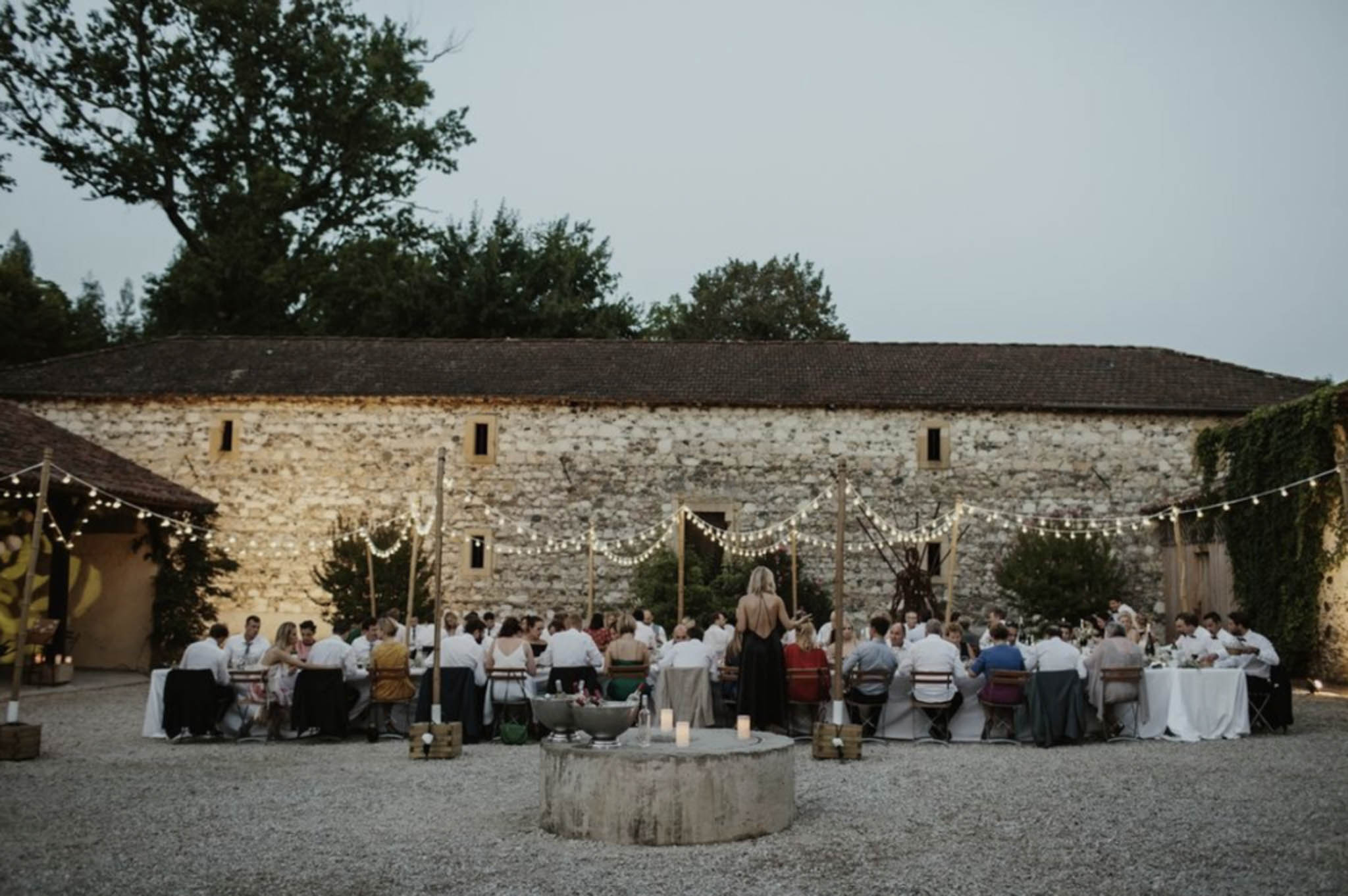 Outdoor wedding reception at dusk in French stone barn courtyard with festoon lights and 50-60 seated guests
