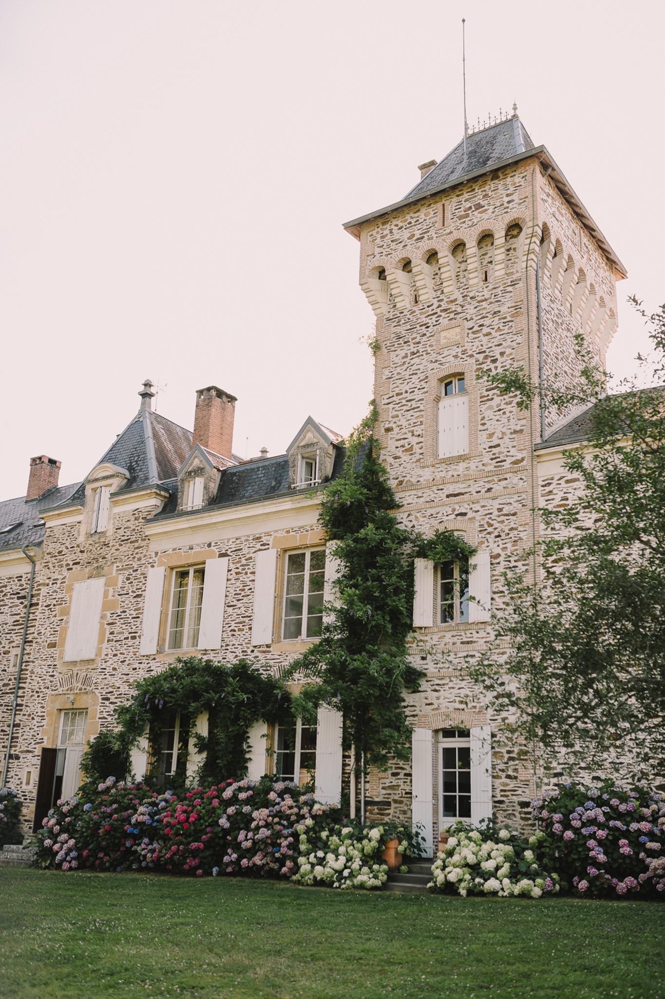 French chateau with square tower, slate roof, white shutters, and pink, white, and blue hydrangea border