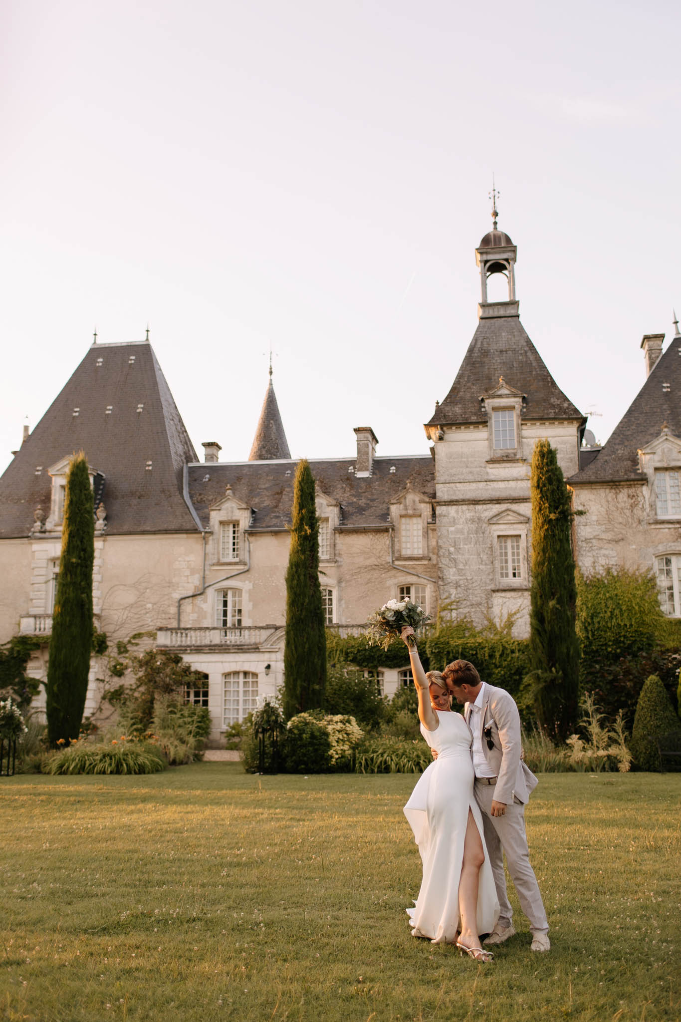 Bride raising bouquet and groom in grey linen suit on chateau lawn with turreted stone facade at golden hour