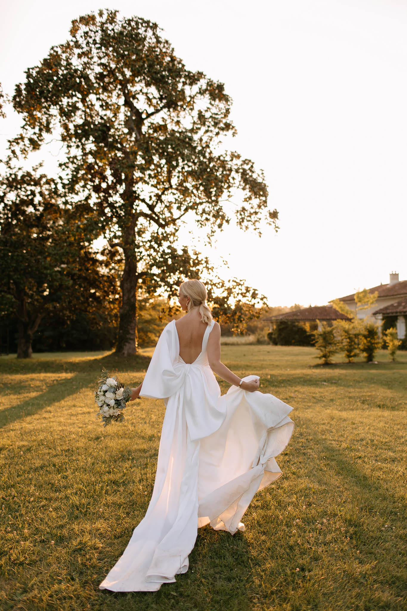 Golden hour rear view of bride walking across estate lawn in open-back gown with bow detail, carrying ivory rose bouquet
