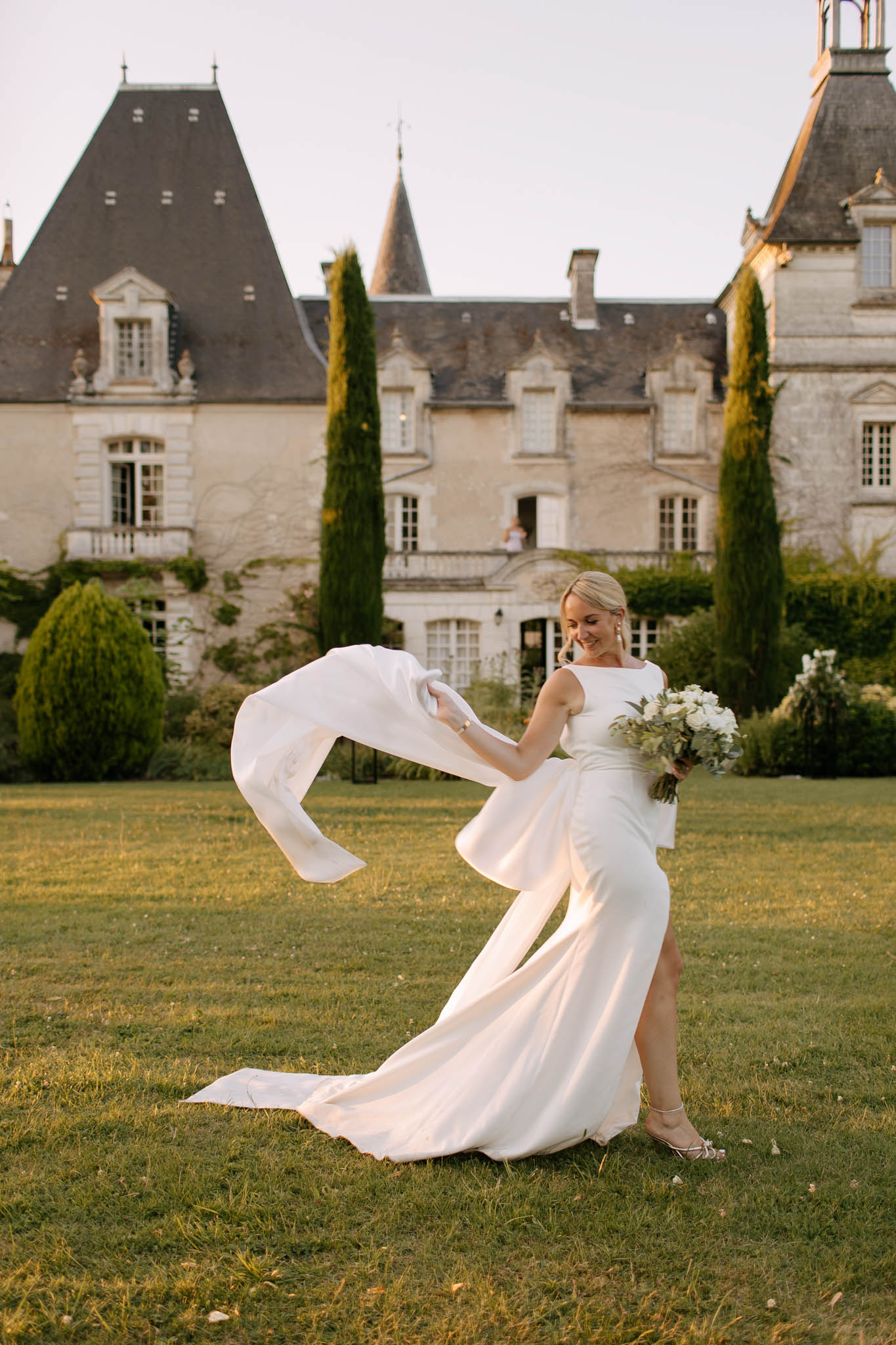 A bride poses alone on the lawn in front of a French château during golden hour, her long ivory veil caught mid-movement as she tosses or adjusts it, creating a sweeping arc of fabric. She wears a sleek, minimalist ivory gown with a bateau neckline, a thigh-high slit, and a train, paired with strappy silver heels and drop earrings. She holds a round bouquet of white roses and green foliage and is smiling downward. The setting is the grounds of a classic French limestone château with steep slate-roofed turrets and tall cypress trees flanking the facade, with a figure faintly visible on the balcony in the background. The composition is a full-length portrait shot in warm late-day light, with a modern, clean styling aesthetic. Potential venue feature image.