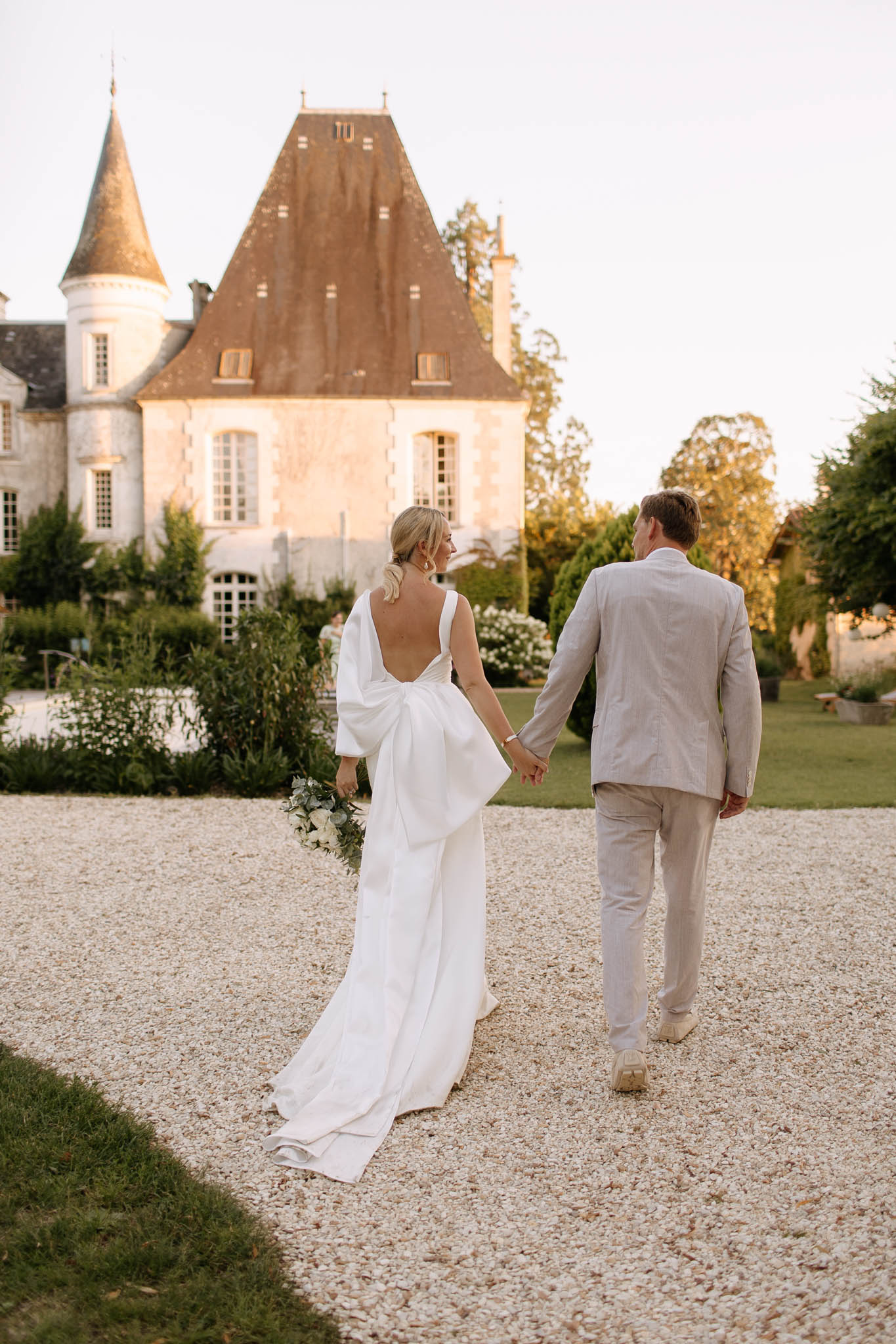 A couple walks hand-in-hand along a gravel path toward a French château during golden hour, photographed from behind in a full-length portrait shot. The bride wears a white satin gown with a deep open back and a large oversized bow detail at the lower back, with a trailing skirt, and carries a small bouquet of white flowers with greenery. The groom wears a light beige linen suit with no tie and casual cream sneakers. The château behind them is a classic French manor with steep terracotta-tiled rooflines, a conical turret, and pale stone façade, surrounded by manicured garden beds with white flowering shrubs. The styling is modern and minimal with a clean, neutral palette. Potential venue feature image.