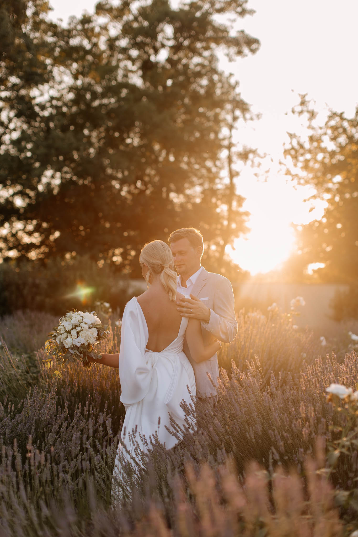 A couple portrait taken outdoors in a lavender field at golden hour, with warm backlit sunlight creating a strong lens flare effect. The bride faces away from the camera, wearing a white open-back gown with draped sleeves and a large bow detail at the lower back; her blonde hair is styled in a low updo. She holds a bouquet of white roses and greenery at her side. The groom, in a light beige suit with a white pocket square and open collar shirt, faces toward the camera with his eyes closed, his hand resting on the bride's shoulder. The foreground and surrounding area feature rows of lavender in bloom, and the background is filled with softly blurred trees illuminated by the setting sun. Medium-distance portrait composition with intentional use of backlight and bokeh.