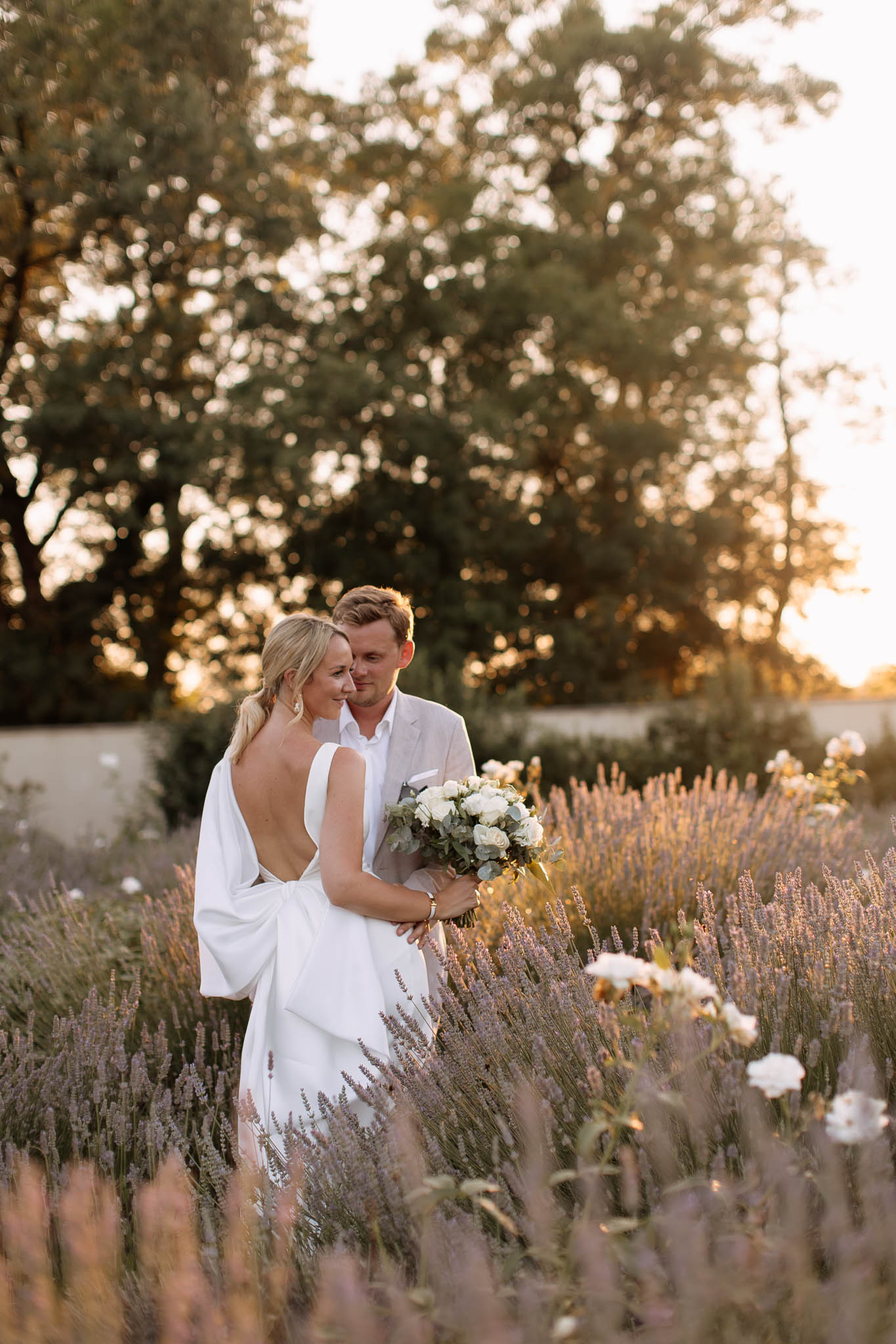 A couple portrait taken outdoors at golden hour in a lavender field, with white roses also visible among the plantings. The bride wears a white gown with a deep open back and a draped bow detail at the lower back, and carries a bouquet of white roses and garden roses with eucalyptus greenery. The groom wears a light beige suit with no tie and a white shirt, standing behind the bride with his arms around her. Both are looking downward with a relaxed, close pose. The composition is a mid-shot taken at ground level, with the lavender framing the foreground and soft bokeh trees in the background. The warm golden-hour backlight gives the image a warm amber tone throughout.