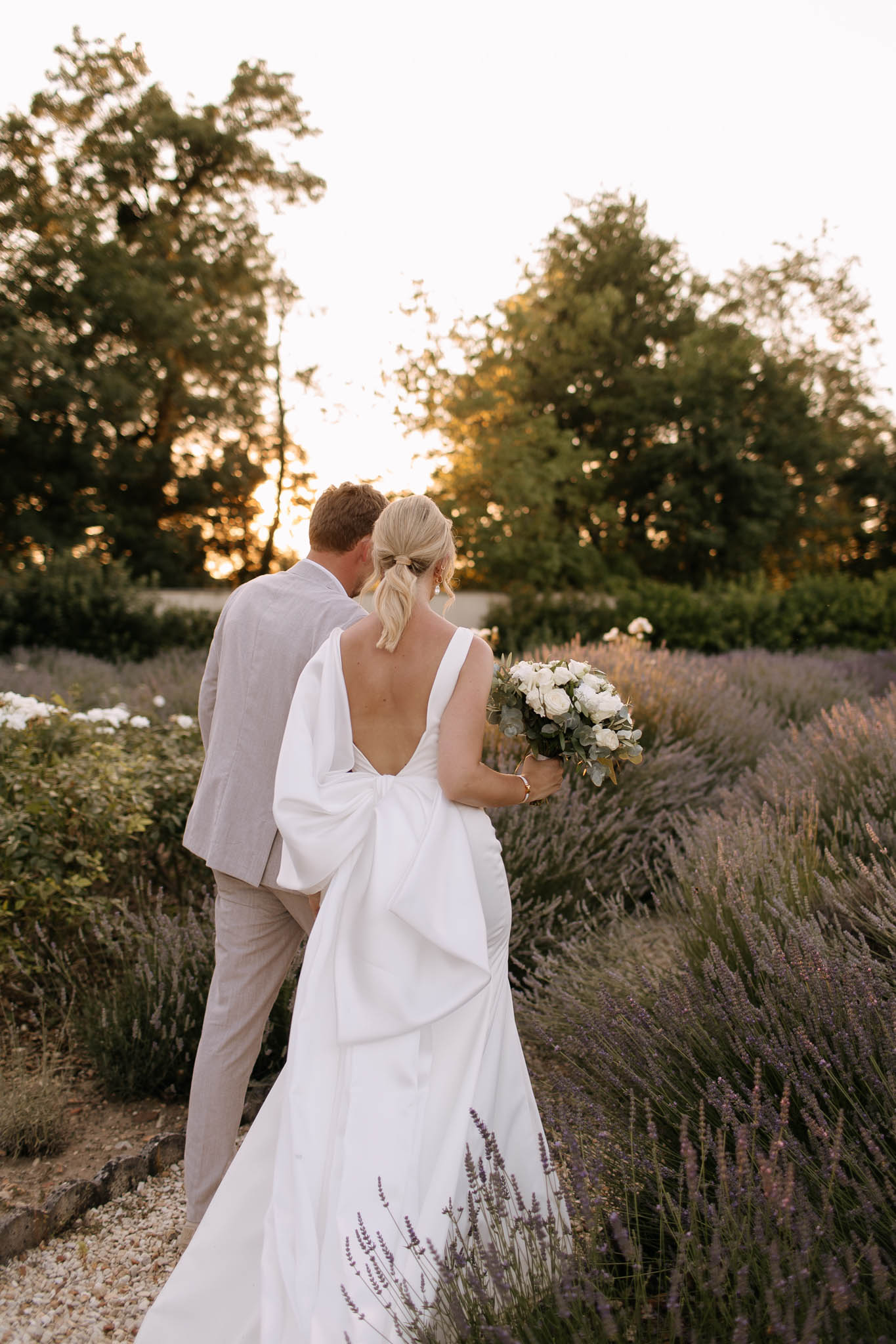 A couple portrait shot from behind, taken outdoors during golden hour in a garden with rows of blooming lavender lining a gravel path. The bride wears a white fitted gown with a deep open back and a large structured bow detail at the lower back, and carries a bouquet of white roses and eucalyptus; her blonde hair is styled in a low ponytail. The groom wears a light beige linen suit. The composition is a medium-distance portrait emphasizing the back details of the bridal gown, with warm golden light filtering through trees in the background.