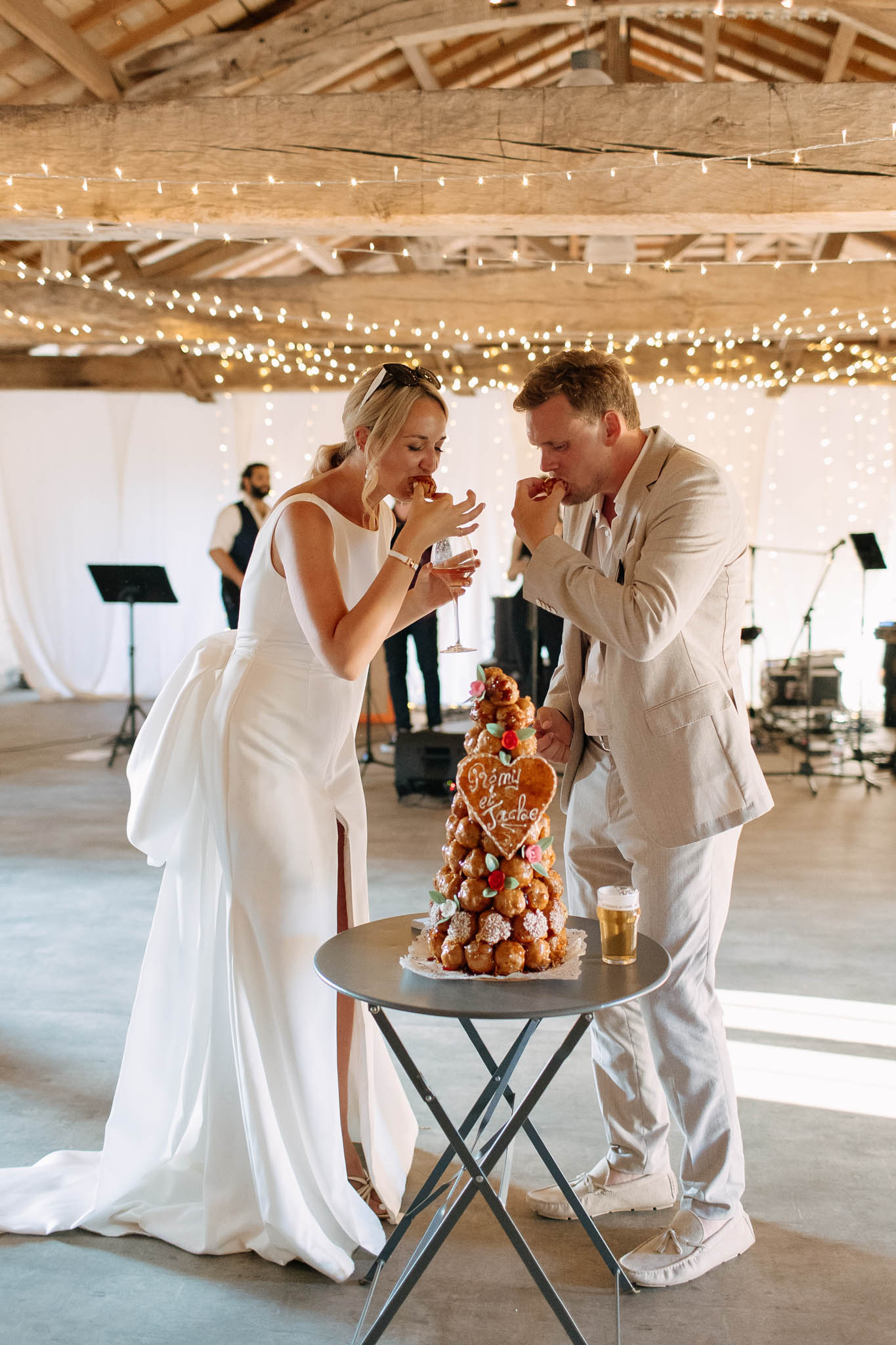 The bride and groom are cutting into and eating from a traditional French croquembouche at their wedding reception, set inside a rustic barn venue with exposed wooden beam ceiling draped in warm fairy lights and white fabric wall draping. The bride wears a sleek white satin gown with a front slit and a train, holding a champagne flute, while the groom is dressed in a light linen suit. The croquembouche is topped with a personalized heart-shaped gingerbread plaque with the couple's names written in icing, decorated with small red and pink sugar flowers, and is displayed on a small round bistro table alongside a glass of beer. A live band musician is visible in the background setting up equipment. The shot is a mid-length portrait capturing the couple's candid reaction as they taste the dessert.