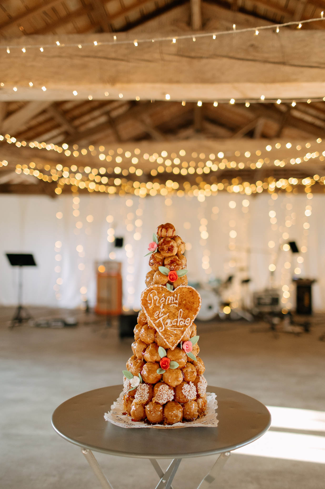 A traditional French croquembouche wedding cake is displayed on a round silver cocktail table in the foreground, decorated with small red sugar roses, green sugar leaves, and a heart-shaped gingerbread plaque reading 'Rémi et Jacbe' in white icing. The profiterole tower is finished with caramel and sits on a white paper doily. The setting is an indoor barn-style reception venue with exposed wooden beam ceiling draped with multiple strands of warm fairy lights that cascade down white curtained walls, creating a rustic-warm atmosphere. A music stand and drum kit are visible in the soft-focus background, indicating a live band setup for the reception. The image is a portrait-format detail shot with the croquembouche sharply in focus against a heavily bokeh background.