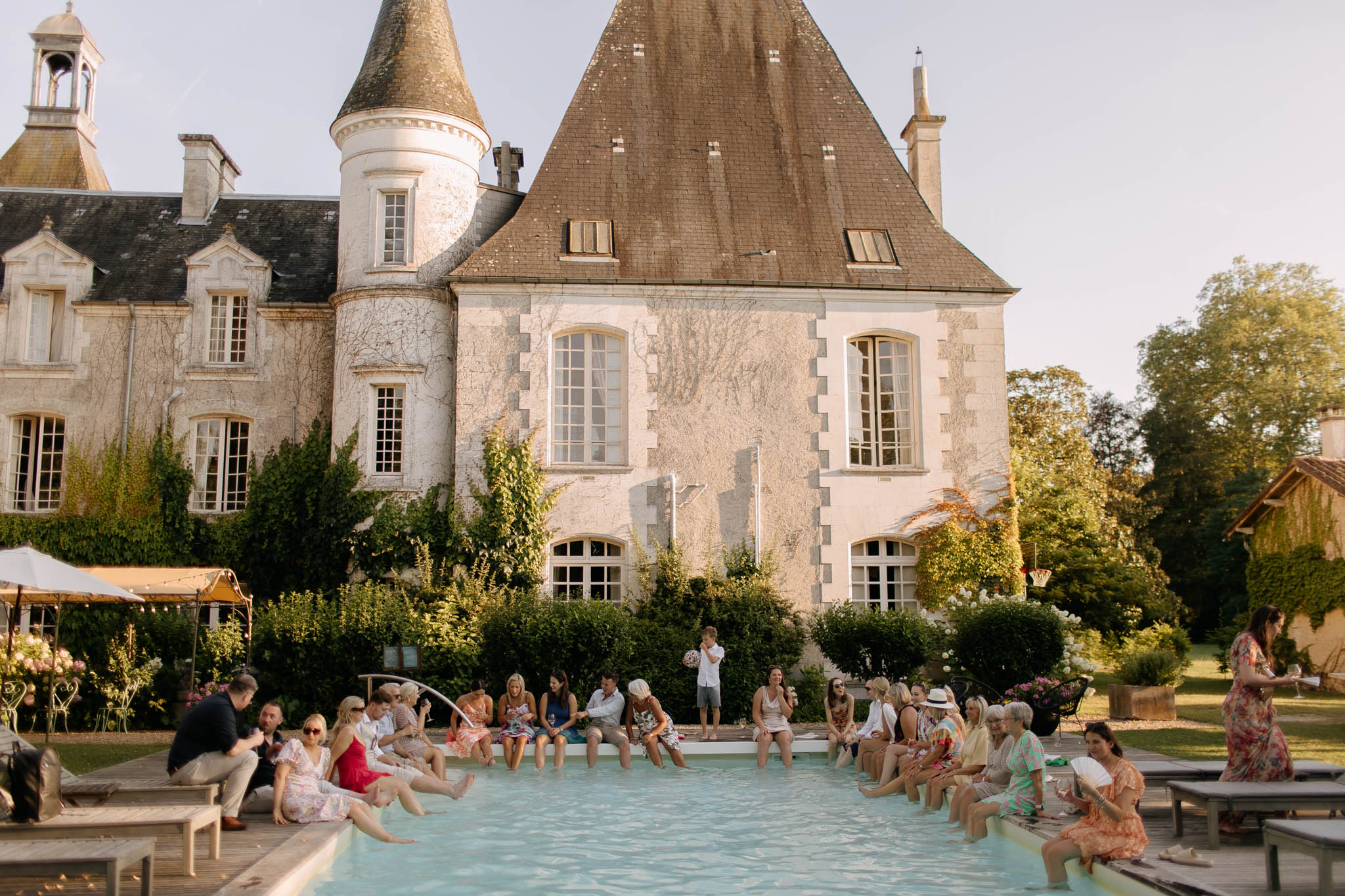 Wedding guests gather poolside during what appears to be a cocktail hour or informal reception moment at a French château. Approximately 25–30 guests are seated along the edge of a rectangular outdoor pool with their feet in the water, socializing and drinking in the warm evening light. The château behind them is a classic French manor with conical-roofed turrets, white limestone facade, and tall arched windows partially covered in climbing vines. Guests are dressed in warm-weather summer attire including floral print dresses, a red dress, and light linens, suggesting a relaxed, garden-party atmosphere. To the left, a canopied area with string lights and a white umbrella is visible alongside floral arrangements in pink tones. The shot is a wide environmental image taken from poolside level, capturing both the crowd and the full façade of the building. Potential venue feature image.