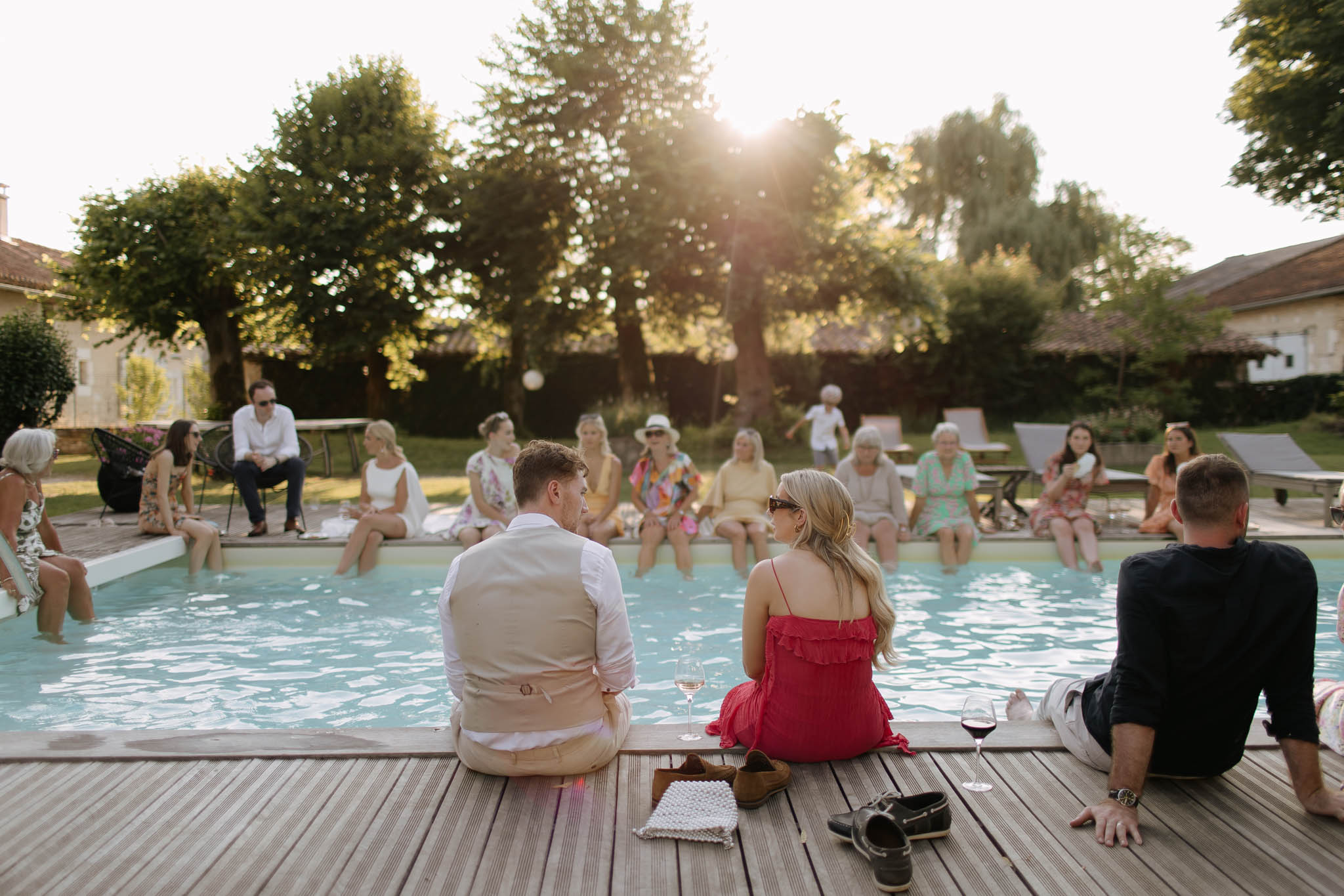 A wedding gathering takes place outdoors around a rectangular swimming pool at golden hour, with approximately 20 guests relaxing poolside at what appears to be a French country estate. In the foreground, a man wearing a beige linen suit and a woman in a red ruffled spaghetti-strap dress sit on a wooden deck with wine glasses beside them — a white wine and a red wine — facing guests seated across the pool with their feet in the water. Guests are dressed in colorful summer attire including a multicolored patterned dress, a yellow dress, a white ensemble, and various bright sundresses, giving the gathering a relaxed, informal feel. The wide shot is taken from behind the foreground couple, with warm backlit sunlight creating a golden flare through the trees, and stone estate buildings are partially visible on either side of the scene.