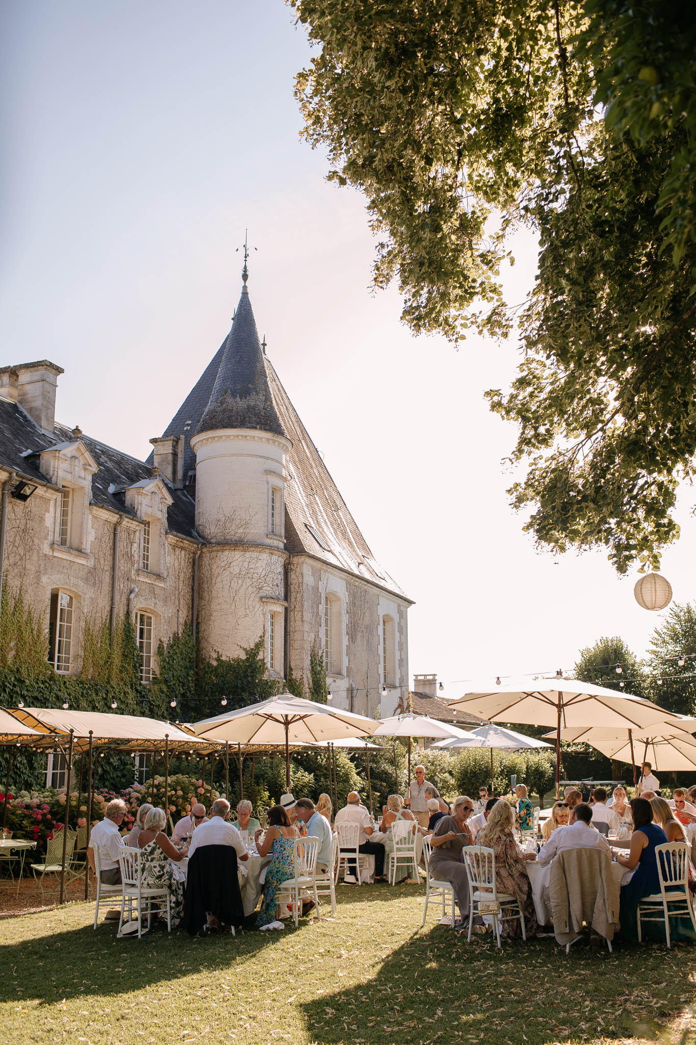 Round tables under cream umbrellas on lawn beside chateau with conical-roofed tower in afternoon sun