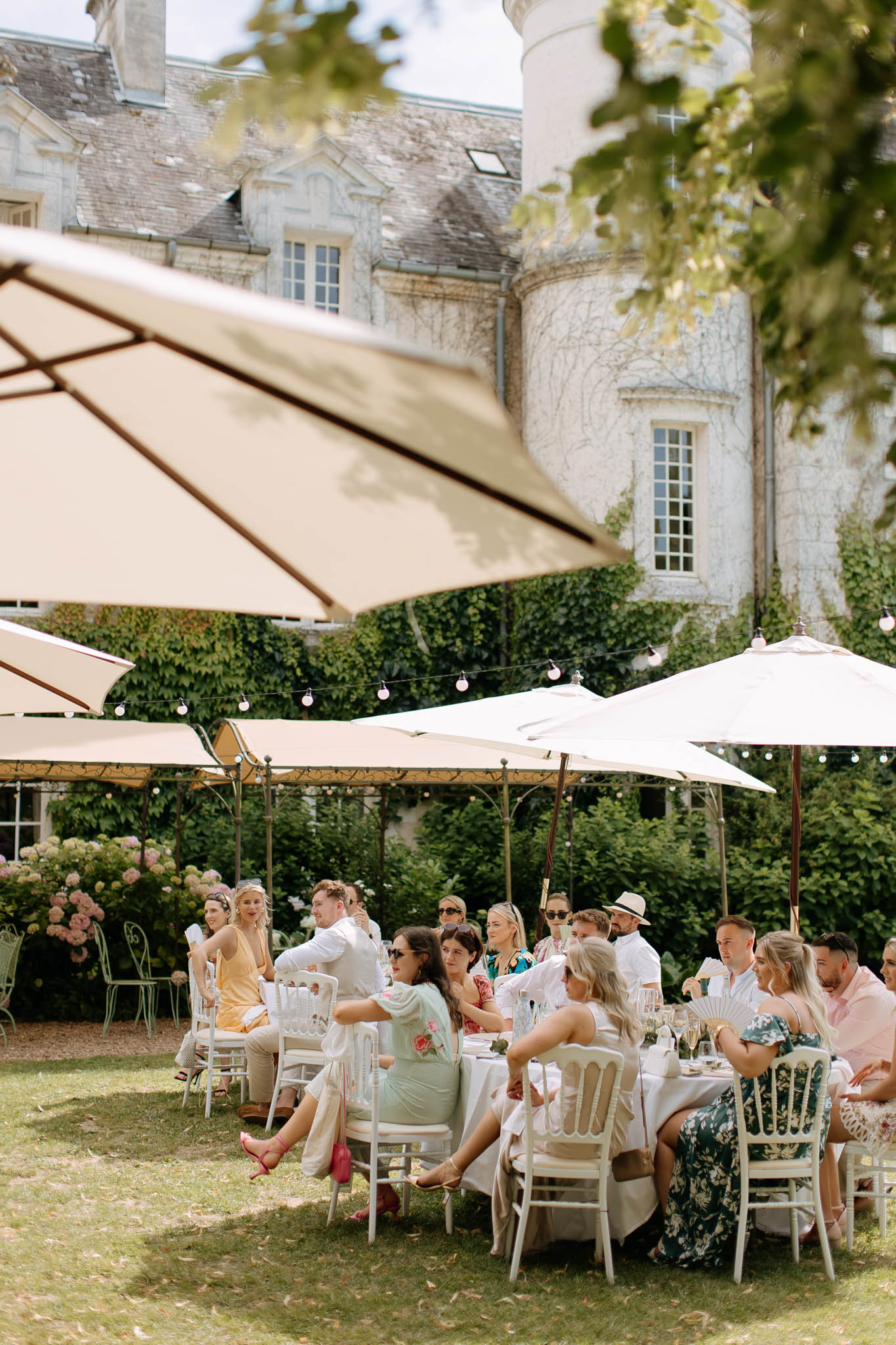 20 guests at round tables with cream parasols on chateau lawn with globe lights and pink hydrangeas