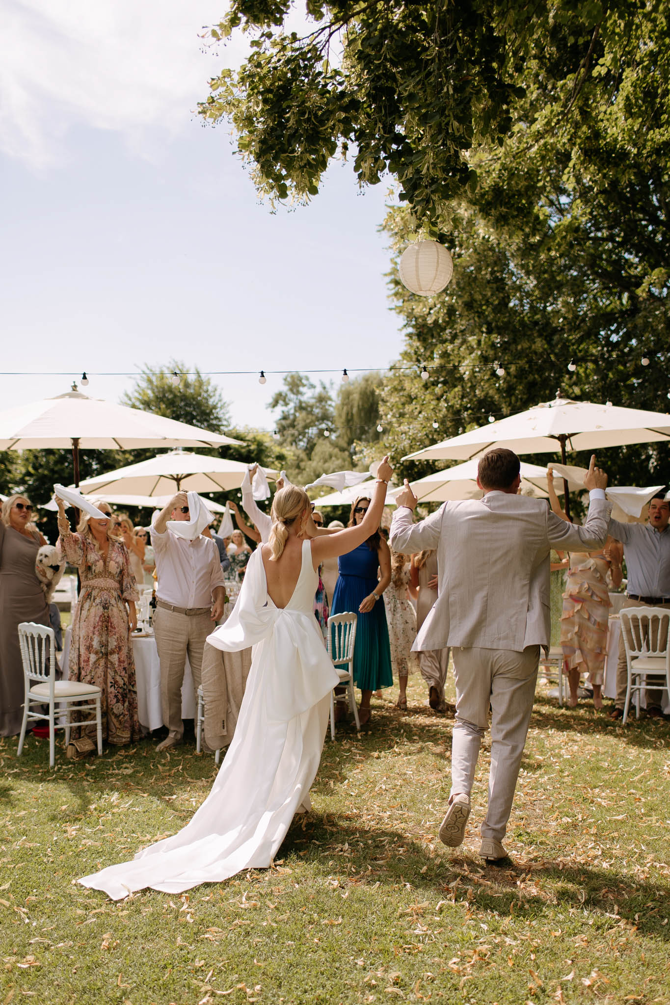 Couple enters garden reception from behind as standing guests wave white napkins under cream parasols