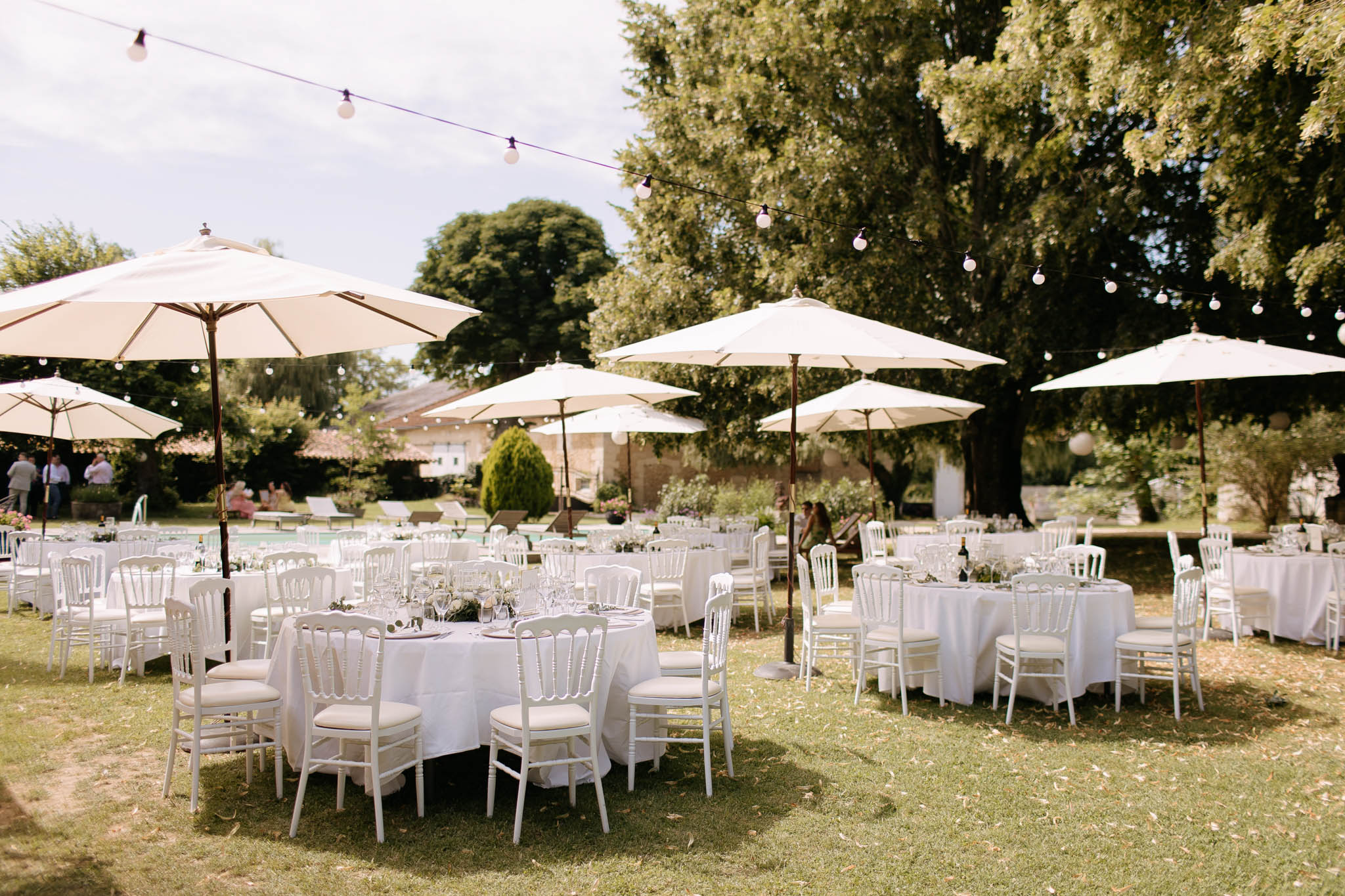 Round white-clothed tables under cream umbrellas with globe lights beside pool at French garden reception