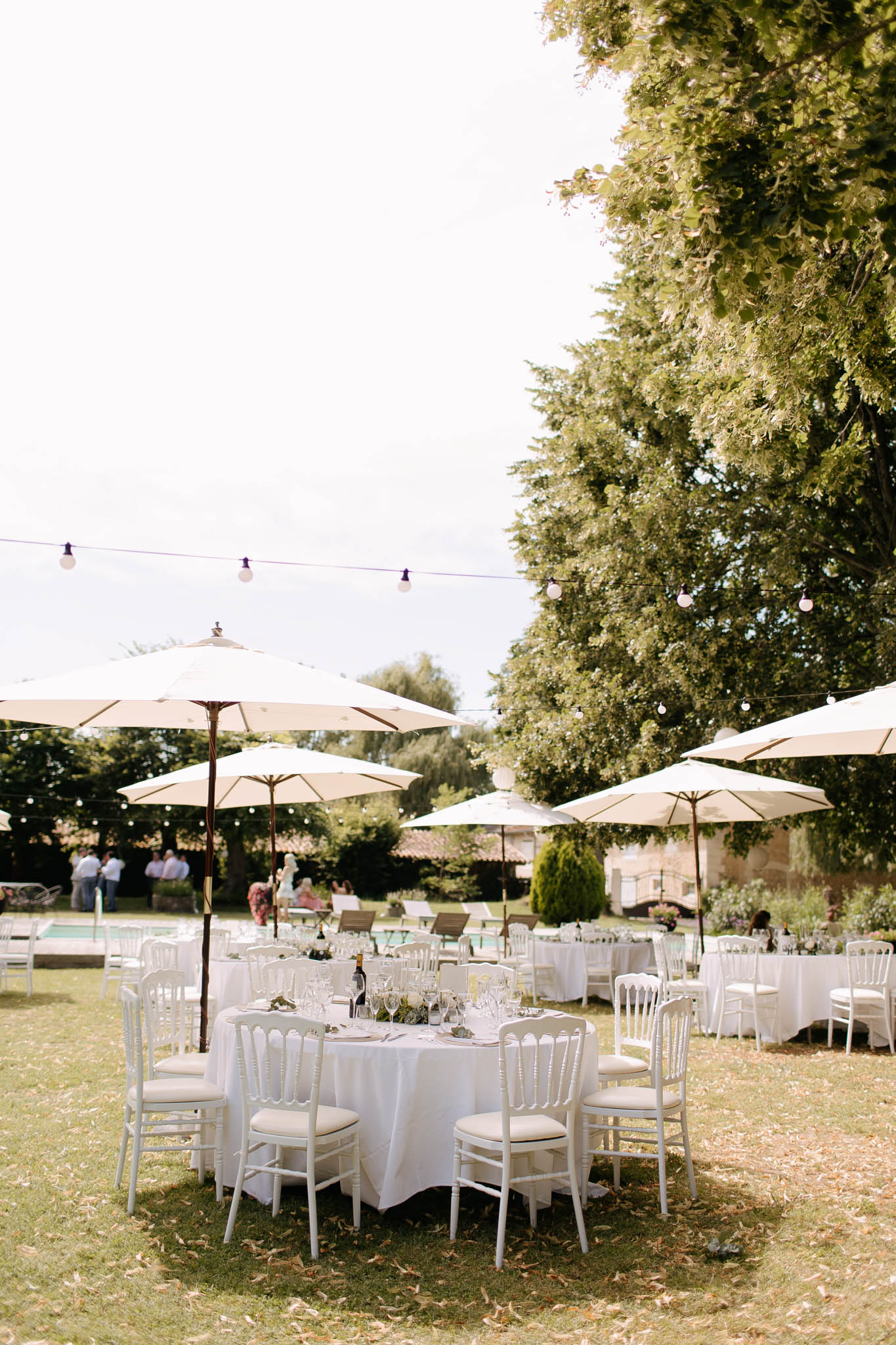 Round tables with white linens and Chiavari chairs under cream umbrellas with fairy lights in garden
