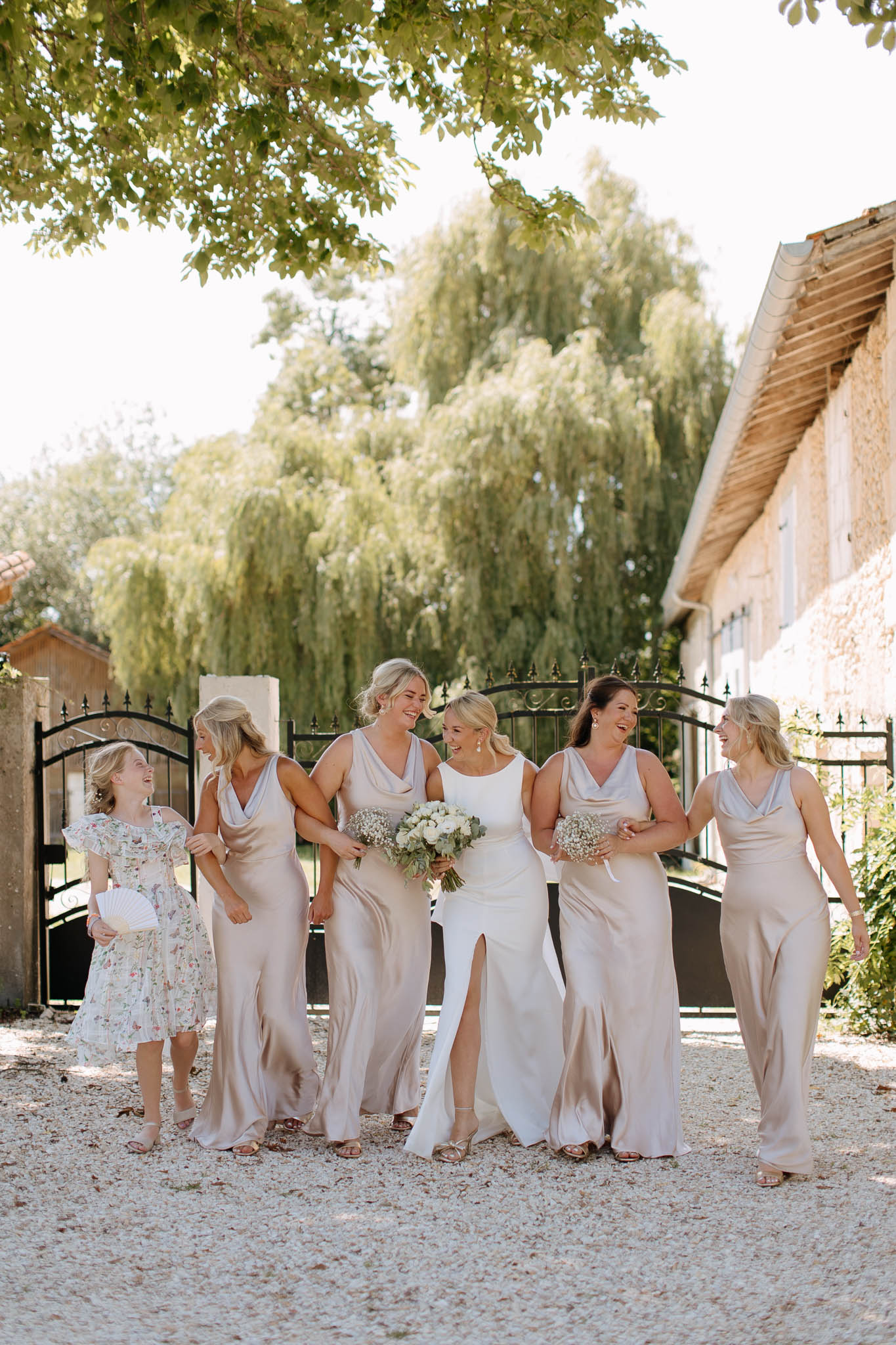 Bride in ivory gown walking with four bridesmaids in champagne satin dresses and flower girl on gravel courtyard