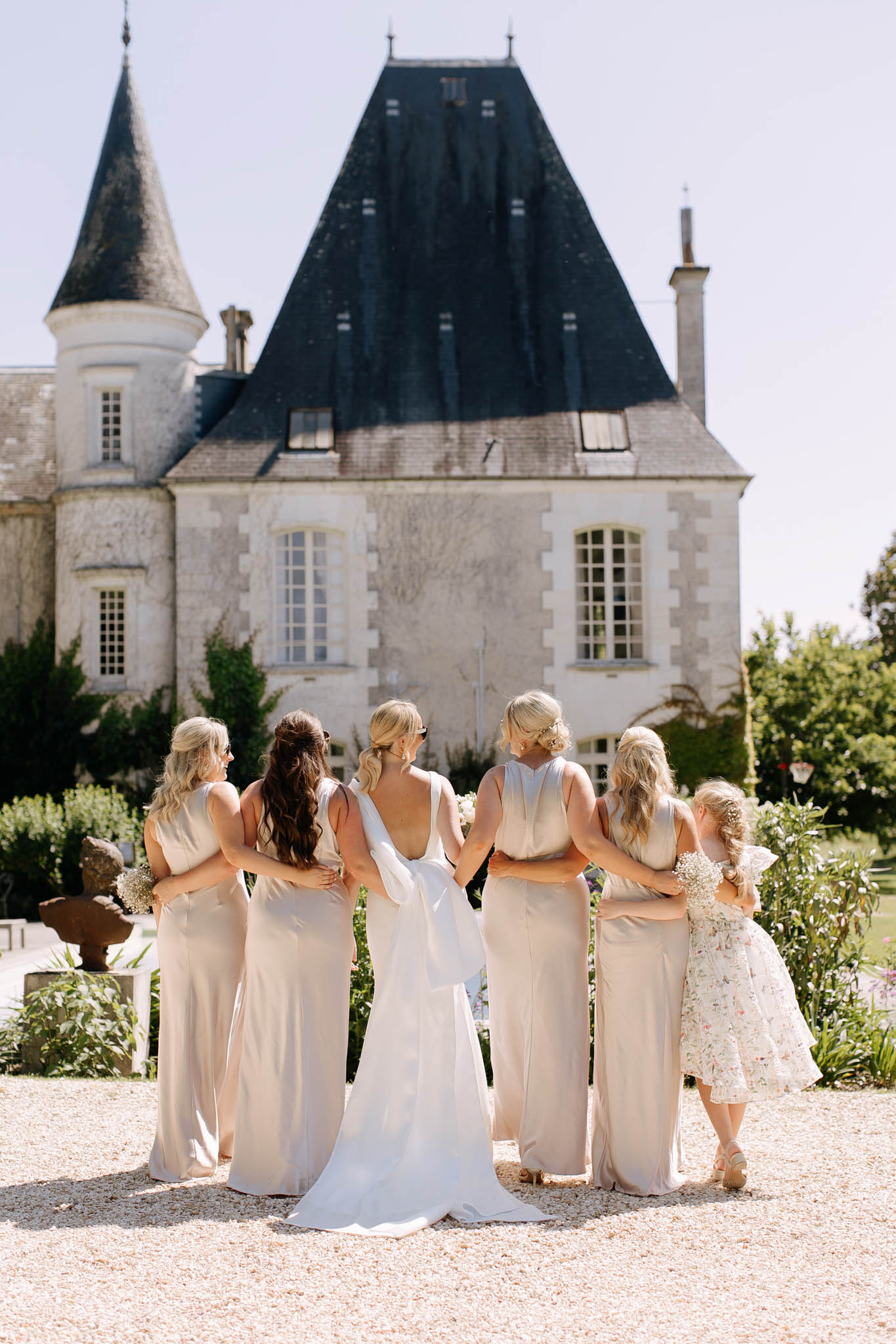 Bride and four bridesmaids in champagne satin dresses stand with backs to camera facing a French chateau