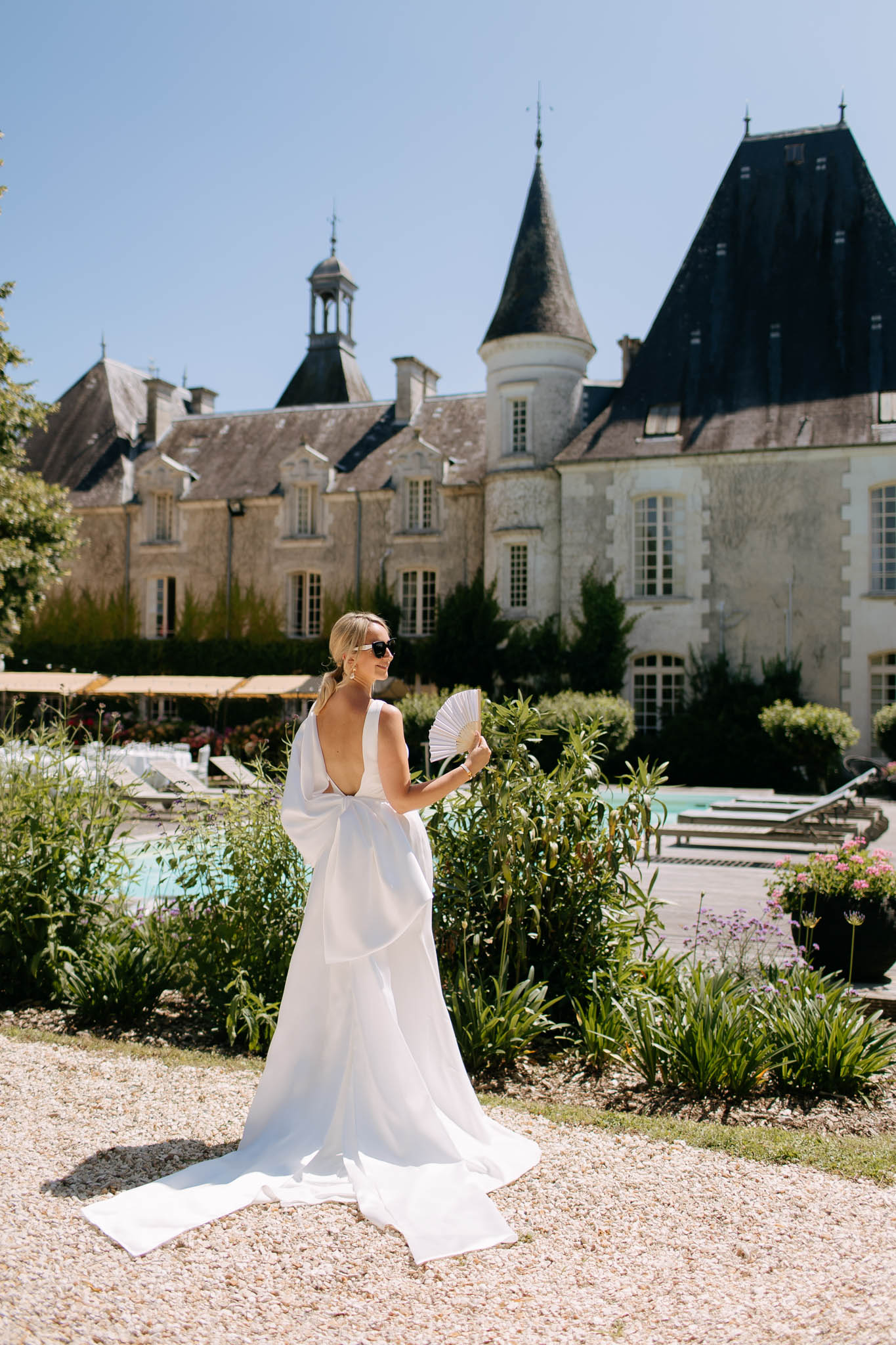 Bride showing deep open-back gown with bow detail holding hand fan with pool and turret chateau behind