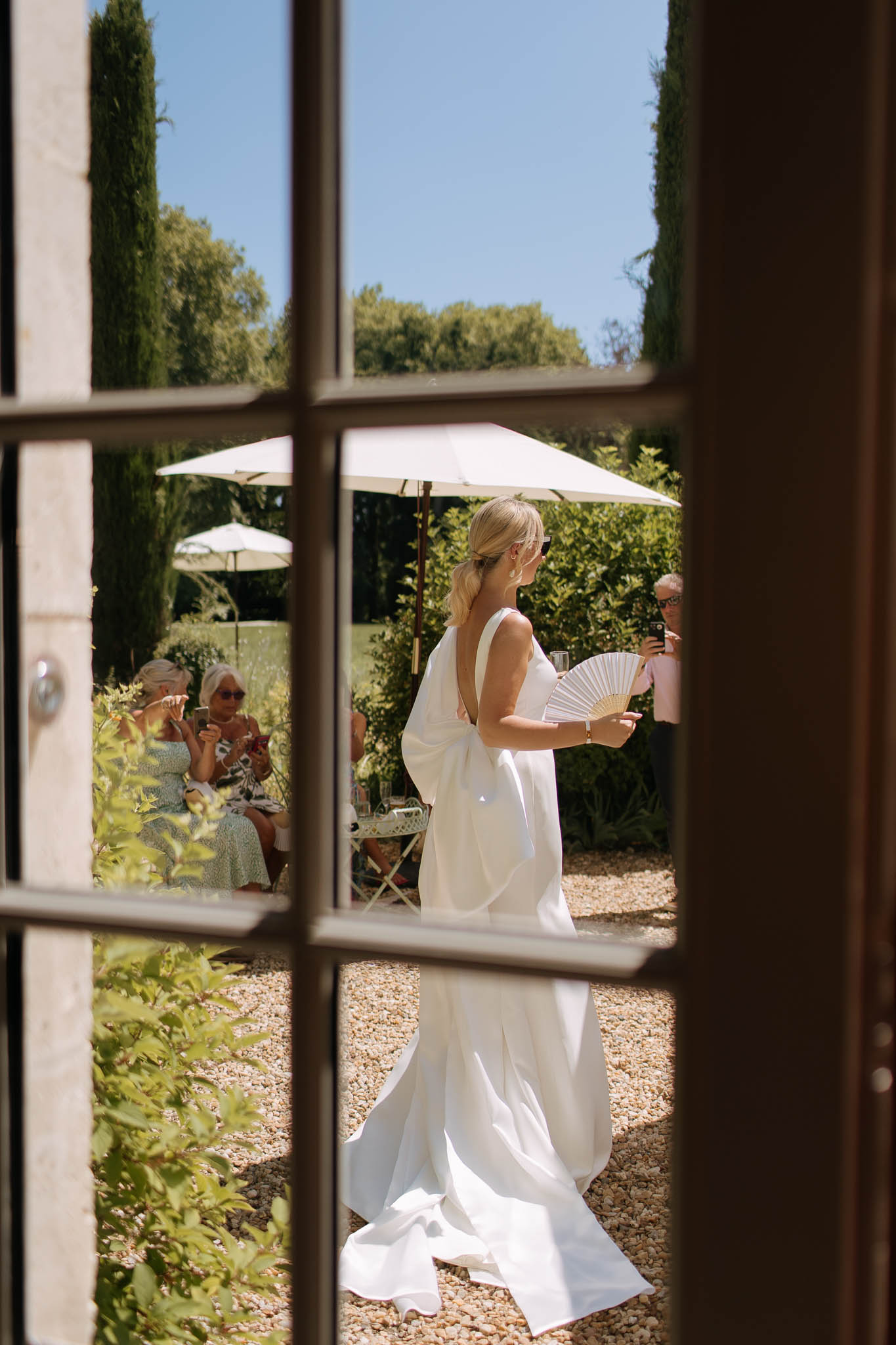 Bride seen from behind through French doors wearing open-back white gown and holding hand fan during cocktail hour