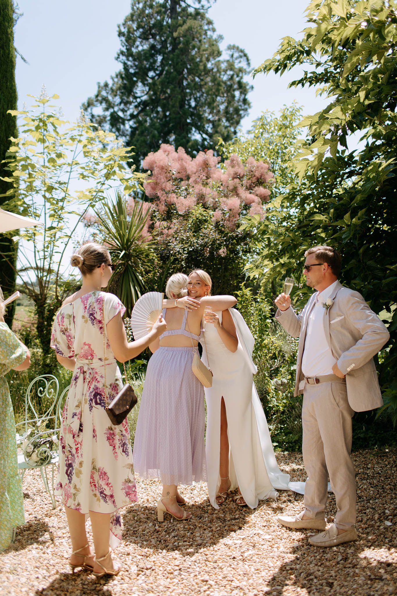 An outdoor cocktail hour scene on a gravel path in a garden setting, where the bride is embracing a female guest. The bride wears a fitted ivory sleeveless gown with a front slit and a long flowing train. The groom stands to the right in a light sand-colored linen suit with a white shirt and a small white boutonniere, holding a champagne flute. A female guest facing away from the camera wears a lavender two-piece crop top and midi skirt with a white handheld fan, while another guest to the left wears a white floral-print midi dress with purple and pink blooms. A partial figure in a sage green outfit is visible at the far left. The group of approximately five people stands on a gravel path surrounded by garden plantings. The overall styling palette is soft and warm — ivory, sand, lavender, and floral prints — consistent with a relaxed, summery garden wedding aesthetic. Medium wide-shot composition.