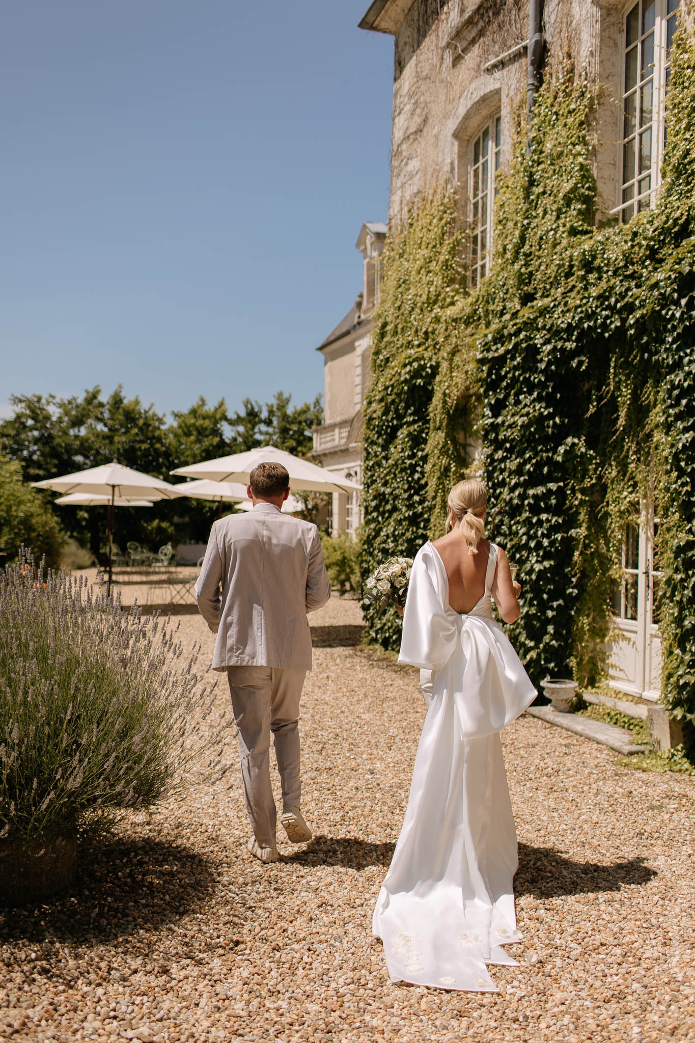 Bride and groom walking from behind along ivy-covered chateau with lavender border and gravel courtyard